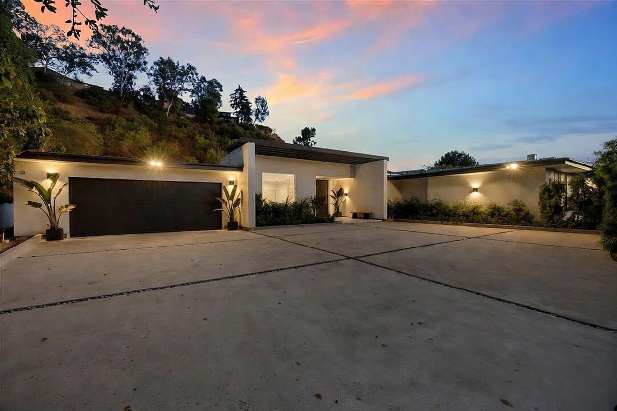 This modern single-story residence features a clean, minimalist architectural style with a flat roofline and a spacious concrete motor court. The exterior is illuminated by warm wall-mounted lighting, highlighting the smooth facade and the dark garage door. Set against a hillside backdrop at dusk, the home presents a sophisticated and inviting curb appeal.