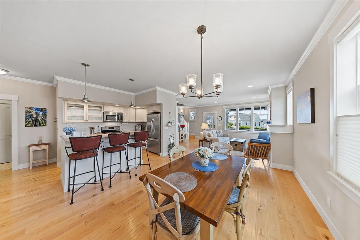 This open-concept living space features a kitchen with a breakfast bar, a dining area, and a glimpse into the adjacent living room. The kitchen is equipped with light-toned cabinetry, stainless steel appliances, and pendant lighting, while the dining area is anchored by a wooden table and chairs. The room is finished with light wood flooring and neutral wall colors, creating a bright and cohesive atmosphere that flows seamlessly between the functional zones.