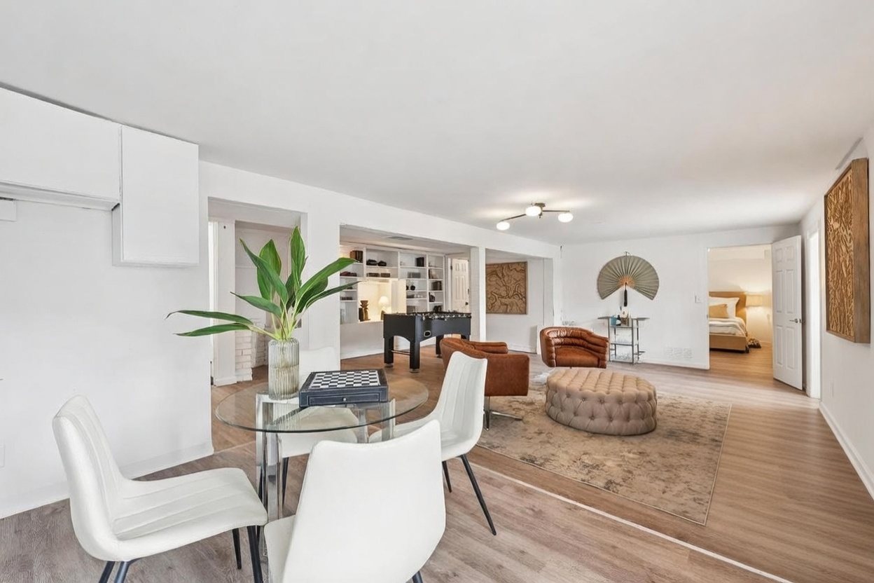 This open-concept living and dining area features light wood-toned flooring and a bright, neutral color palette. The space includes a dining area with a glass-topped table, a central living zone with a tufted ottoman and leather armchairs, and a rear alcove housing a foosball table and built-in shelving. The layout is spacious and airy, with a cohesive design that flows seamlessly between the dining and lounge areas.