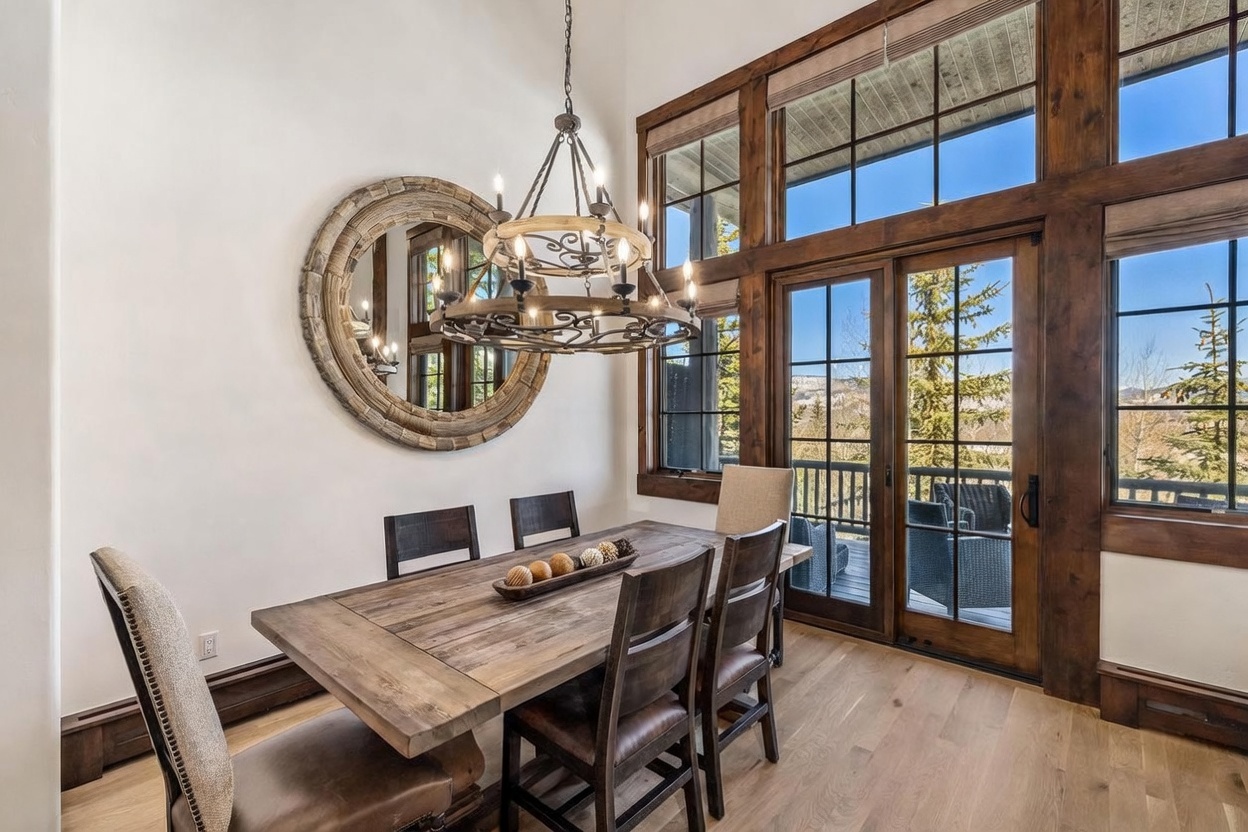 This rustic-inspired dining room features a substantial wood table centered beneath a large, multi-tiered iron chandelier. A prominent circular mirror with a textured frame hangs on the wall, reflecting the space, while tall windows with dark wood trim provide an expansive view of the outdoors. The room is finished with light wood flooring and a mix of upholstered and wooden chairs, creating a warm and inviting atmosphere.
