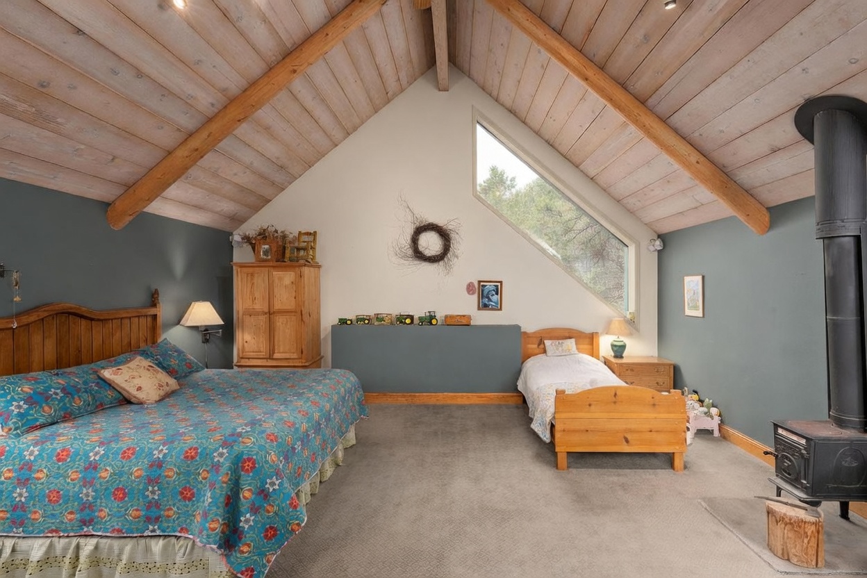 This spacious bedroom features a vaulted ceiling with exposed wooden beams and a large, angled window that brings in natural light. The room is furnished with both a large bed and a smaller twin bed, creating a versatile sleeping area, and includes a wood-burning stove on a hearth. The decor has a rustic, cozy aesthetic with blue-toned walls and light wood furniture.