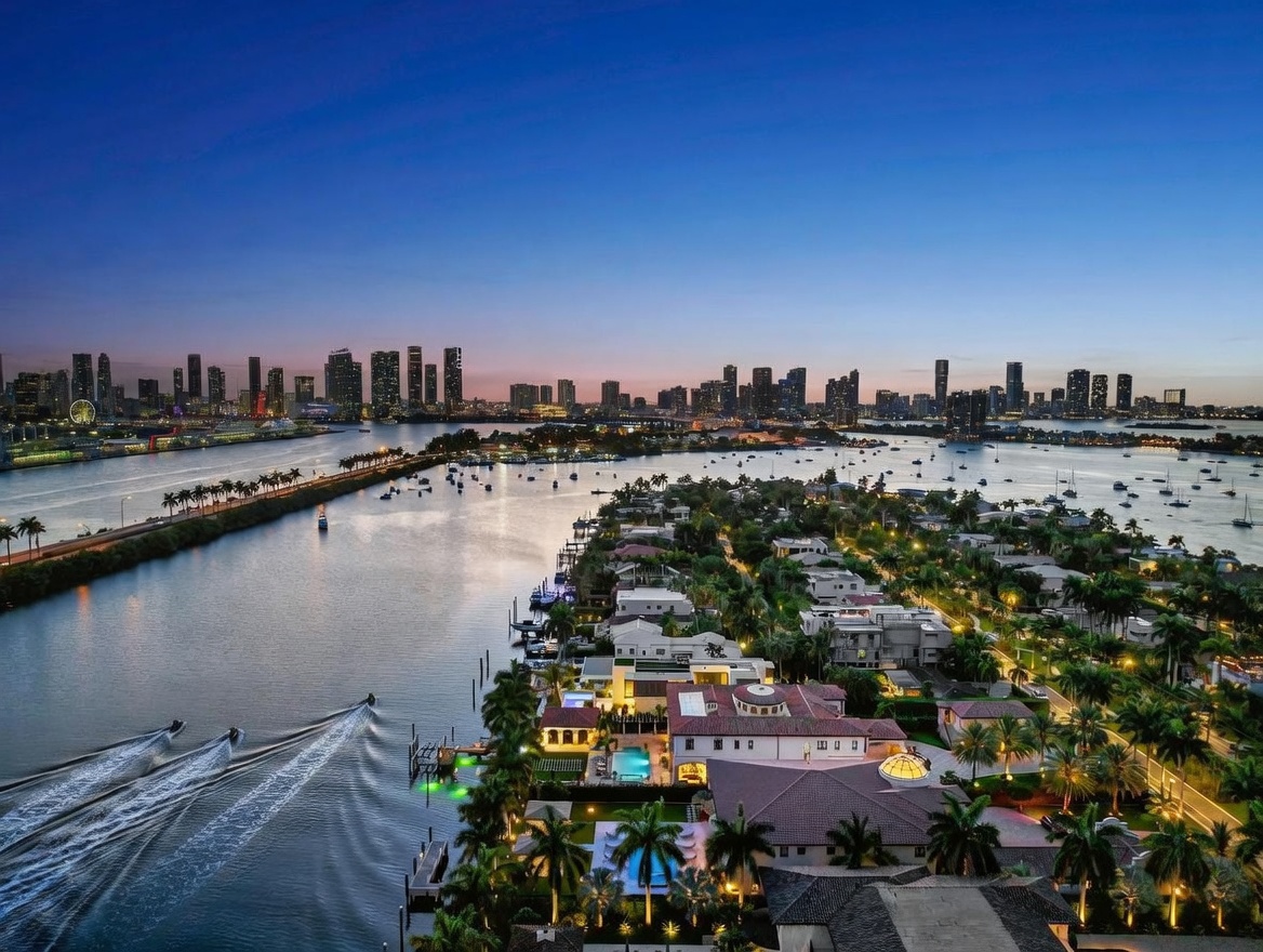 This stunning aerial view captures a luxurious waterfront estate at twilight, showcasing its expansive property, private swimming pool, and direct access to the waterway. The scene features a panoramic perspective of the surrounding neighborhood and the distant city skyline, emphasizing the prime location and serene atmosphere. The warm glow of the home's lighting contrasts beautifully with the cool blue tones of the evening sky and the calm water.