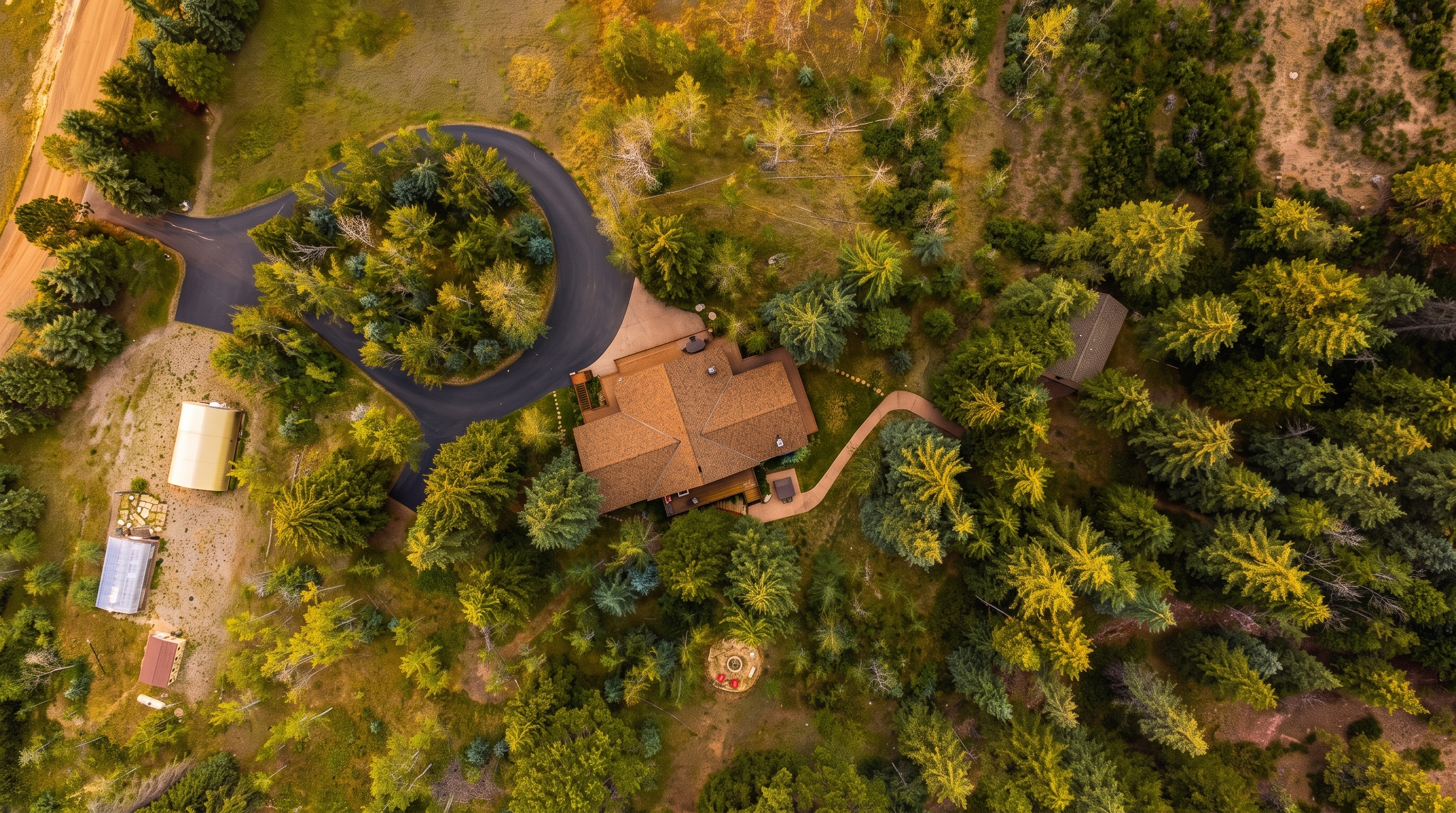 This high-angle aerial perspective captures a secluded residential property nestled within a dense, wooded landscape. The home features a multi-faceted roofline and is accessed by a curved paved driveway, with additional outbuildings and a small fire pit area visible on the surrounding grounds. The setting offers a sense of privacy and immersion in nature, highlighting the expansive, tree-filled lot.