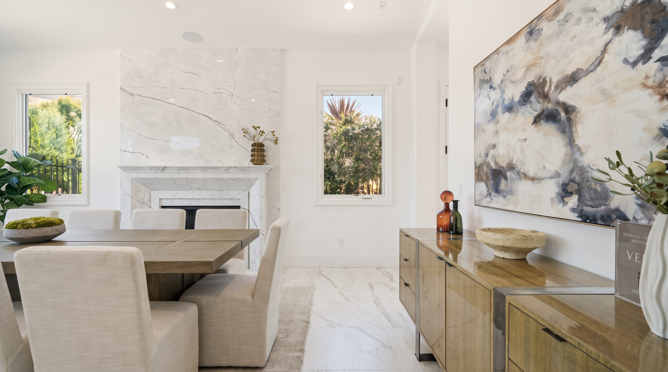 This elegant dining room features a large wooden table surrounded by light-colored upholstered chairs, set against a backdrop of bright white walls and polished stone flooring. A prominent stone-clad chimney breast serves as a focal point, while a sleek sideboard sits to the right beneath a large, abstract piece of wall art. The space is bathed in natural light from two windows, creating an airy and sophisticated atmosphere.