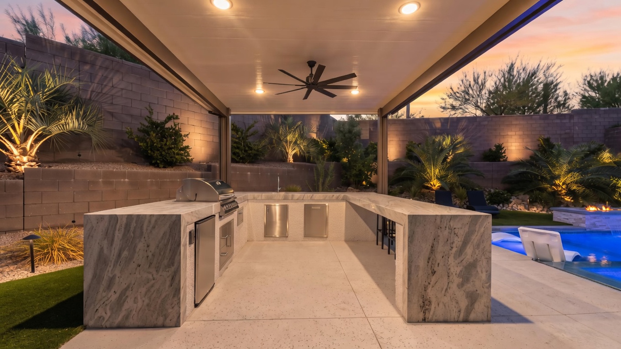 This outdoor kitchen area features a U-shaped stone-topped island with a built-in grill, stainless steel cabinets, and integrated seating. The space is covered by a modern patio roof equipped with recessed lighting and a ceiling fan, creating a comfortable transition to the adjacent swimming pool. The setting is illuminated by warm landscape lighting, highlighting the stone wall backdrop and the surrounding desert-style landscaping.