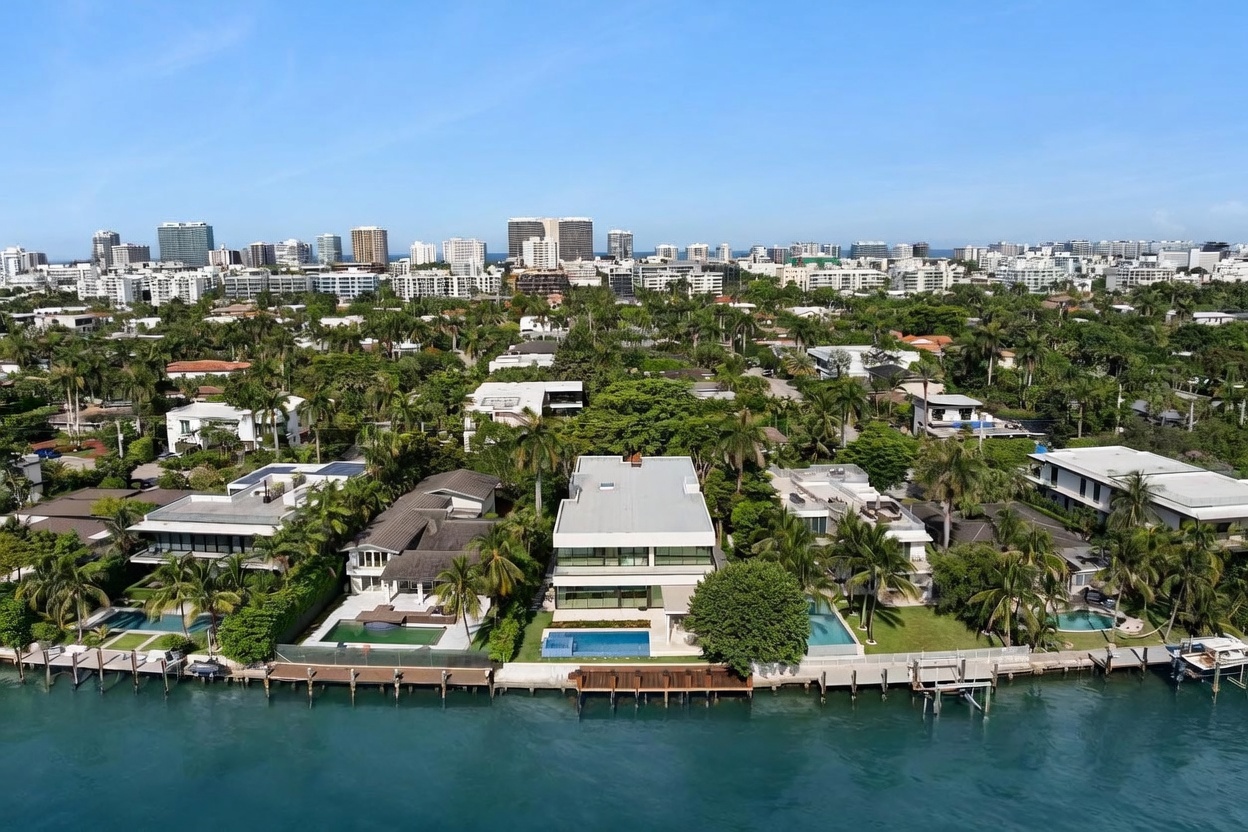 This high-angle aerial perspective captures a row of luxury waterfront residences along a calm canal, showcasing their private docks, swimming pools, and lush tropical landscaping. The central focus is a modern, multi-level home with clean lines and expansive glass, set against a backdrop of a dense residential neighborhood and a distant city skyline. The bright, clear daylight emphasizes the vibrant blue of the water and the well-manicured green lawns of the properties.