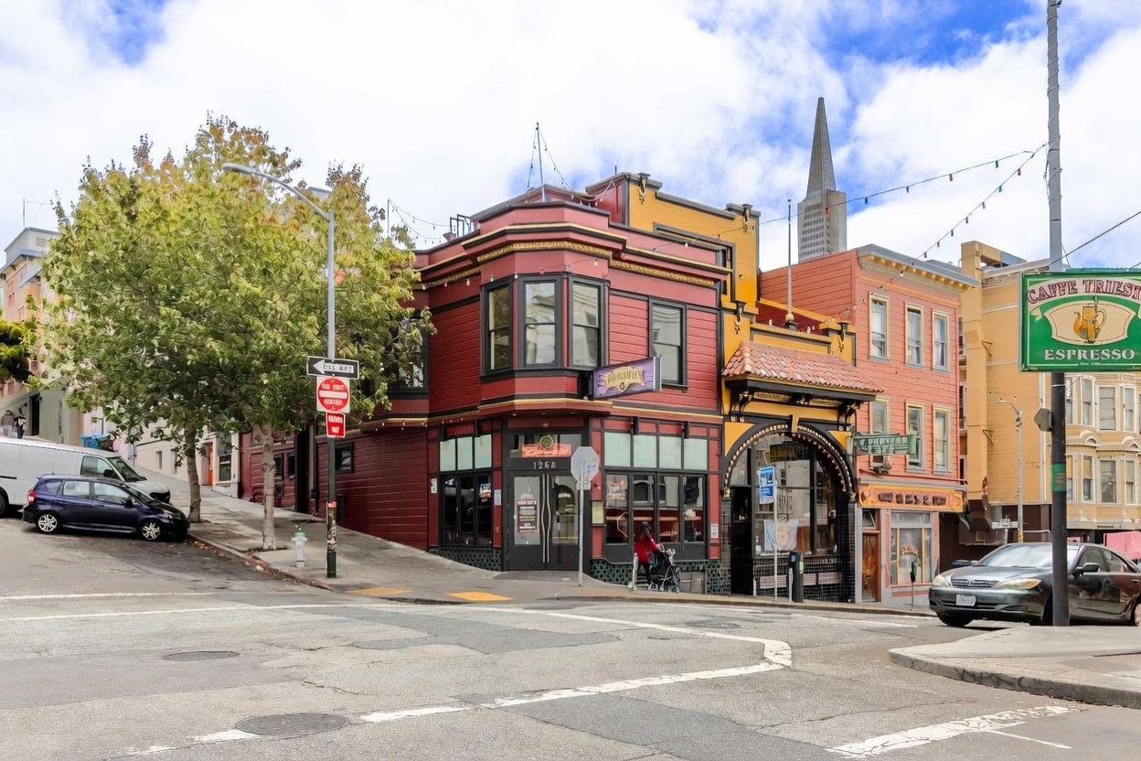 This street-level view captures a distinctive corner building in an urban setting, featuring a red-painted facade with dark trim and large storefront windows. The structure displays classic architectural details, including a prominent bay window section and an arched entryway leading to an adjacent storefront. The scene is set on a city street corner with a clear view of the surrounding neighborhood, including a distant skyscraper and a prominent green sign for Caffe Trieste.