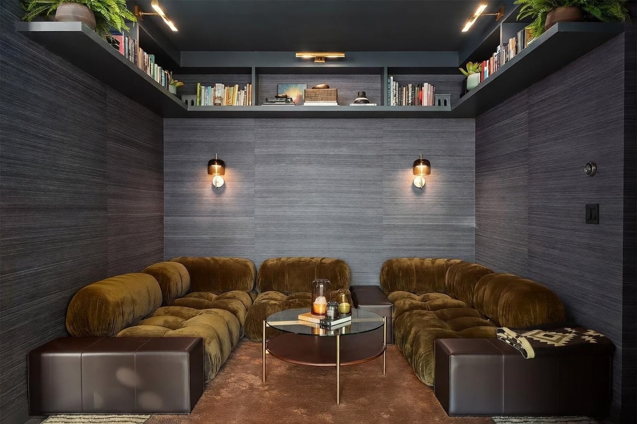 This cozy, moody lounge area features a deep, modular velvet sofa in a rich olive tone, arranged in a U-shape around a circular glass-topped coffee table. The walls are covered in a dark, textured horizontal-patterned wallpaper, complemented by built-in shelving that spans the upper perimeter of the room, accented by integrated lighting. Two warm wall sconces provide soft illumination, creating an intimate and sophisticated atmosphere perfect for relaxation.