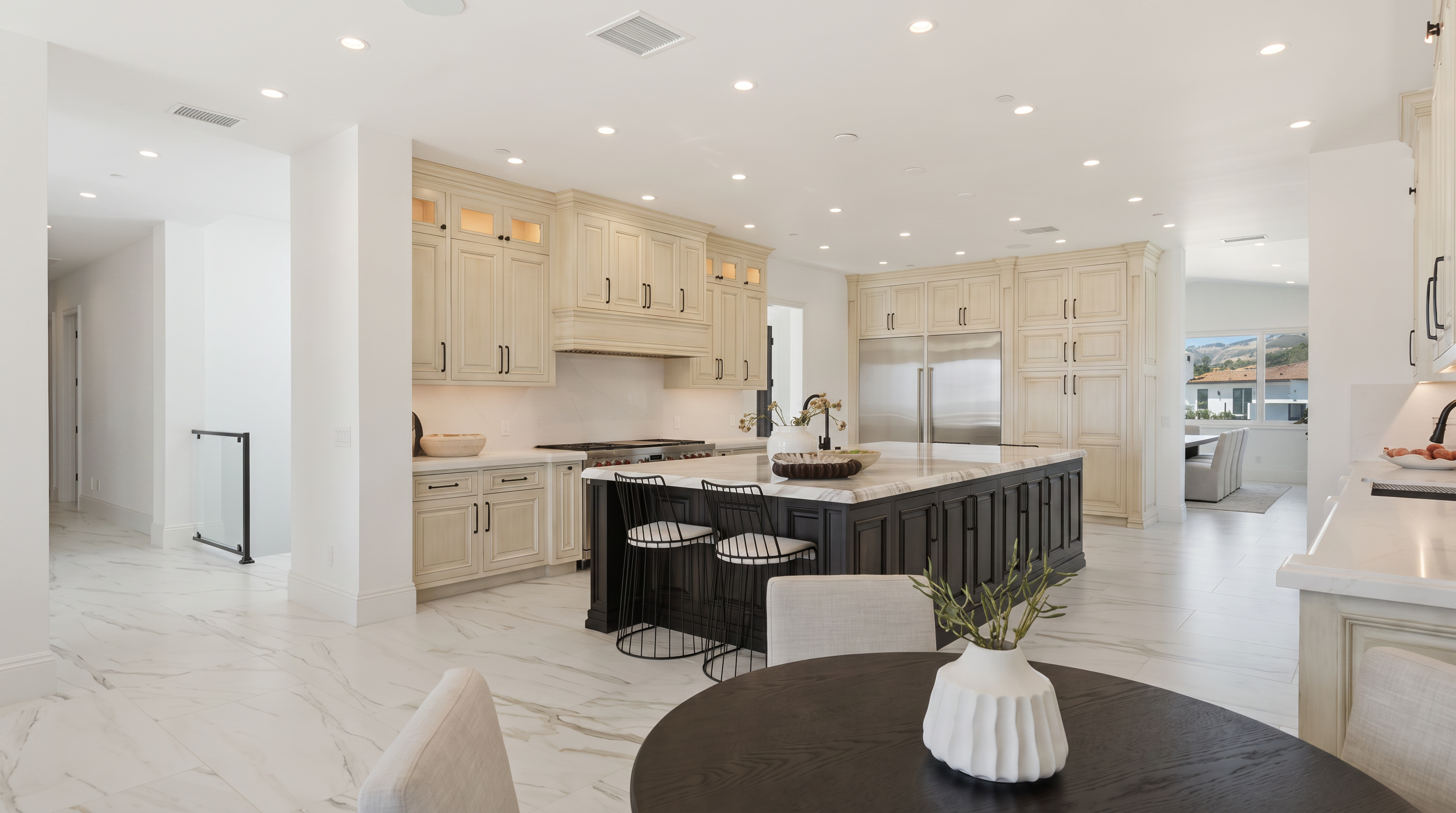 This spacious, open-concept kitchen features a large central island with a dark base and a light stone countertop, complemented by light-toned cabinetry and high-end stainless steel appliances. A round dining table sits in the foreground, creating a seamless transition between the cooking and eating areas. The room is brightened by numerous recessed lights and large windows, showcasing a clean, modern aesthetic with polished light-colored flooring throughout.