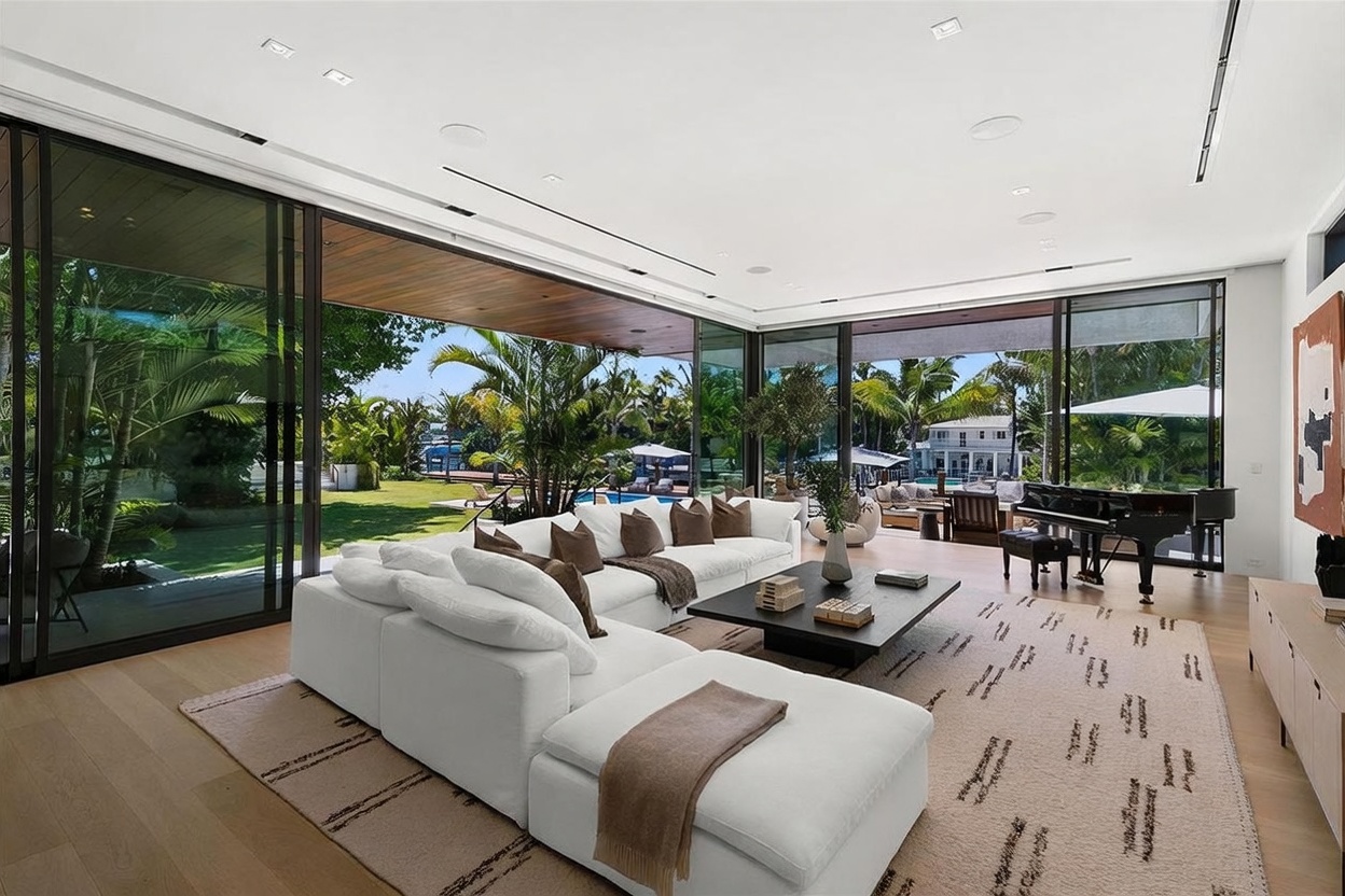 This expansive, modern living room features a large white sectional sofa and a central coffee table set atop a textured area rug. The space is defined by floor-to-ceiling glass walls that slide open to reveal a lush backyard with a pool, creating a seamless indoor-outdoor living experience. A grand piano sits in the corner, and the room is finished with light wood flooring and recessed lighting, offering a bright and airy atmosphere.