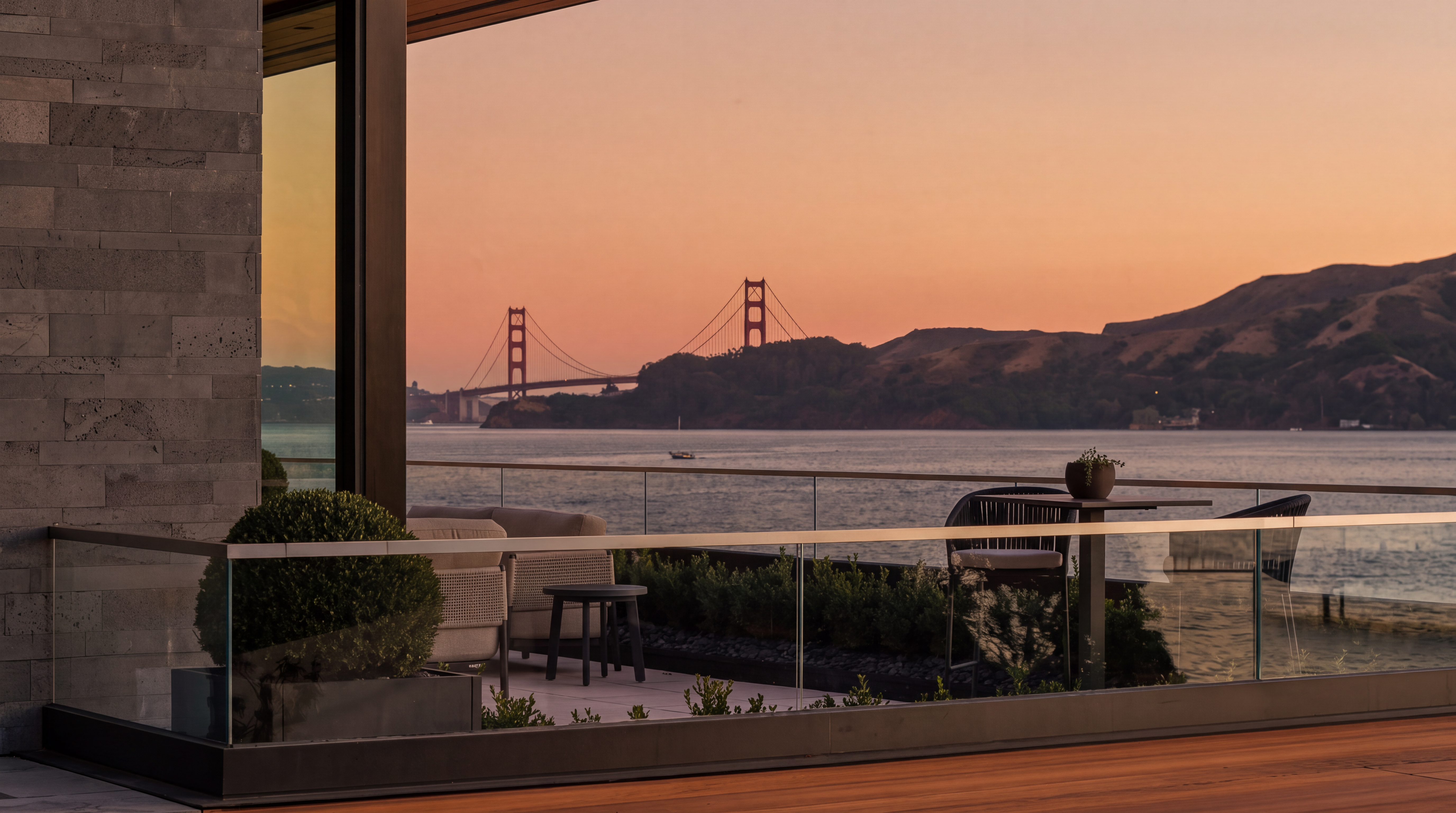 This sophisticated balcony offers a breathtaking, unobstructed view of the Golden Gate Bridge at sunset, framed by the structure's modern architectural lines. The outdoor space is furnished with contemporary seating and a small bistro table, creating an intimate setting for enjoying the panoramic waterfront scenery. The seamless glass railing ensures that the dramatic horizon remains the focal point of this luxurious terrace.