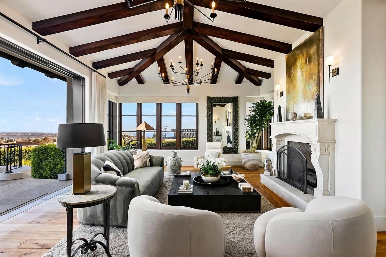 This sophisticated living room features a vaulted ceiling with exposed dark wood beams and a large, ornate stone fireplace as a central focal point. The space is furnished with a plush, curved sofa, two rounded accent chairs, and a substantial dark wood coffee table, all arranged on a textured area rug. Large windows provide abundant natural light and offer expansive views of the surrounding landscape, creating an airy and elegant atmosphere.