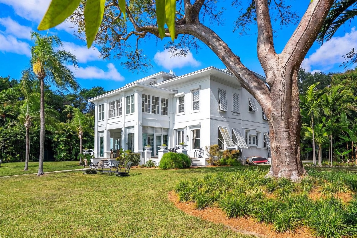 This side view of a large, white, multi-story colonial-style home showcases its classic architectural details, including shuttered windows and a wrap-around porch area. The house is nestled in a lush, green landscape with mature trees and manicured lawn, creating a serene and private atmosphere. A small patio area with outdoor seating is visible near the base of the home, emphasizing the property's potential for outdoor living.