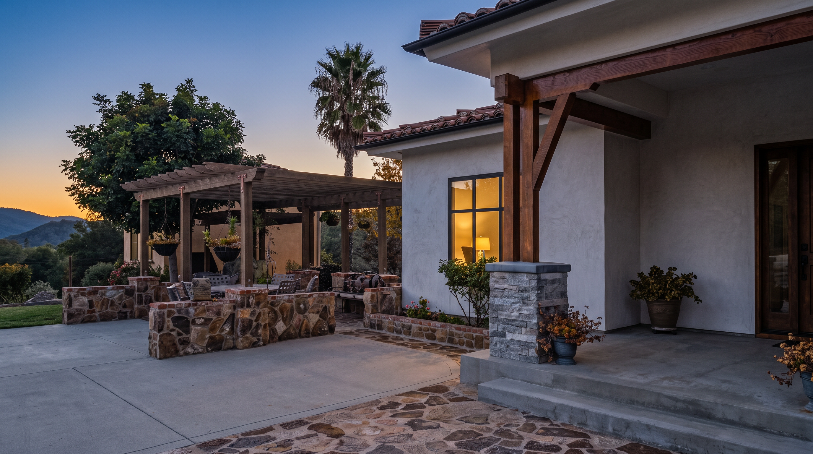 This outdoor living space features a charming stone-accented patio and a wooden pergola covering a comfortable seating area. The home exhibits a clean, light-colored stucco exterior with a tiled roof, accented by decorative stone masonry on the columns. The overall design provides an inviting and sophisticated Mediterranean-inspired atmosphere, perfect for outdoor relaxation or entertaining.