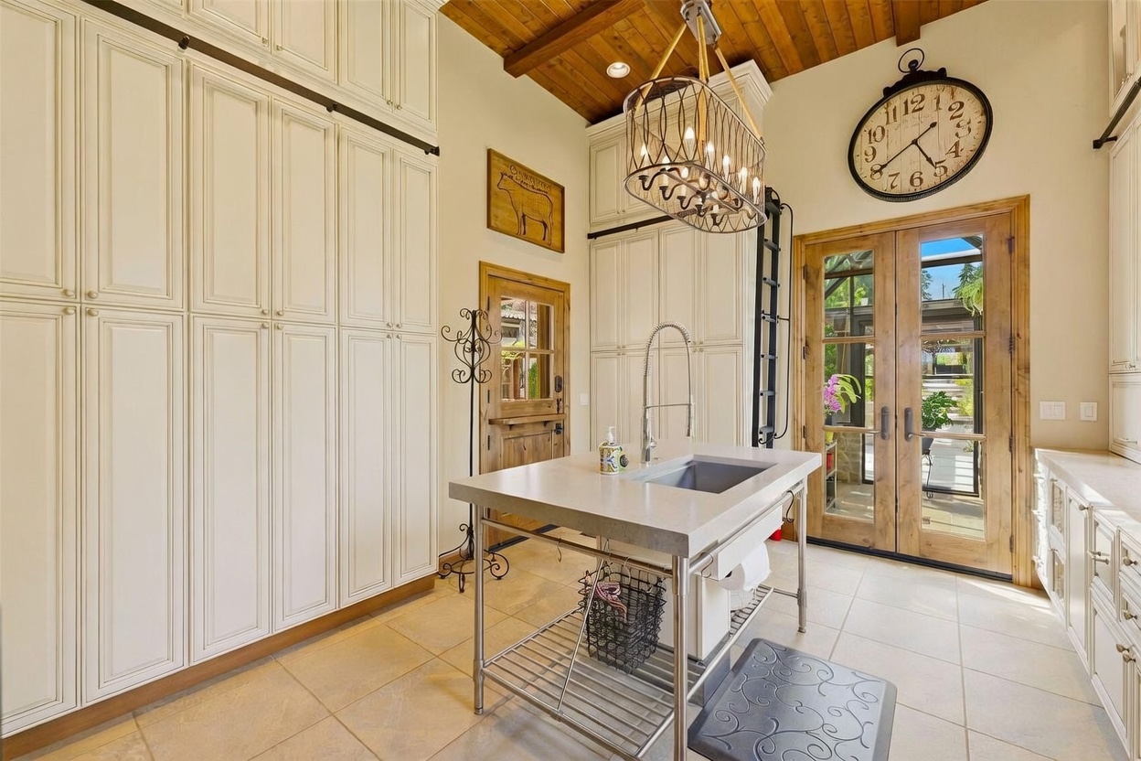 This kitchen features a unique, rustic-industrial aesthetic with a central metal-framed island topped with a stone surface and an integrated sink. The space is defined by tall, cream-colored cabinetry with sliding ladder hardware, a wood-plank ceiling, and a large decorative wall clock above a set of glass-paned double doors. A distinctive cage-style chandelier hangs over the island, adding a touch of character to this well-lit, functional culinary space.