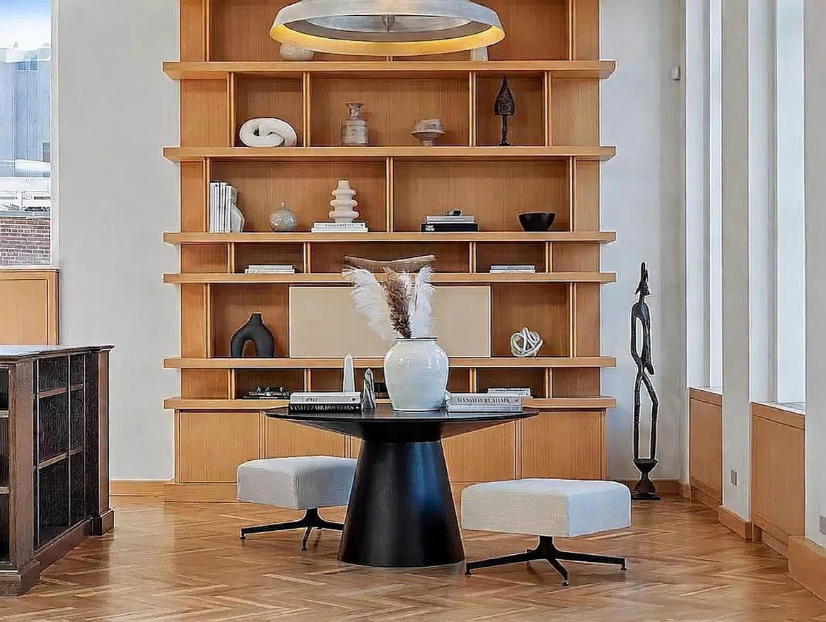 This sophisticated space features a large, light wood shelving unit that serves as a focal point, filled with curated decorative objects and books. A sleek, black pedestal table sits in the foreground, flanked by two modern, light-colored upholstered chairs, creating an intimate seating area. The room is bathed in natural light from tall windows on the right, highlighting the warm tones of the wood flooring and the minimalist, elegant aesthetic.