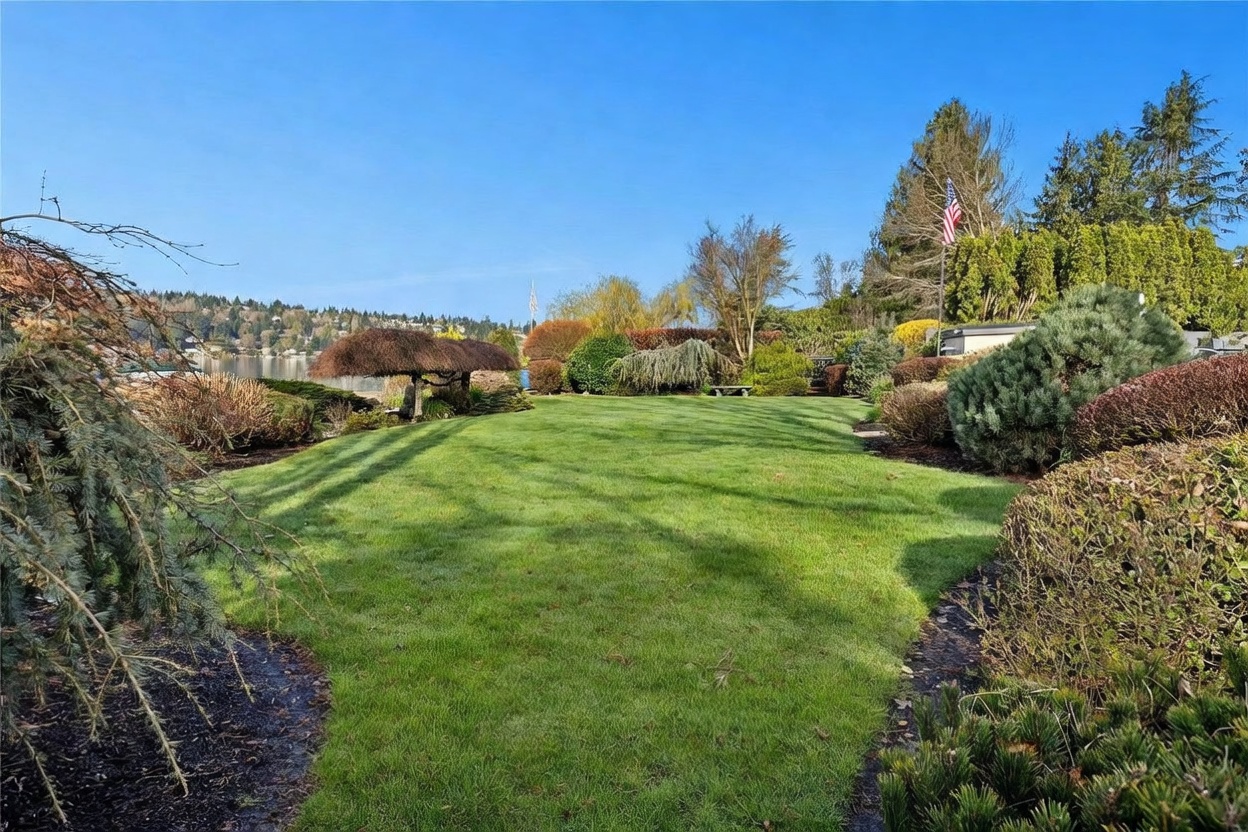This expansive, meticulously manicured lawn serves as a serene backyard retreat, framed by lush evergreen trees and mature landscaping. The perspective looks out toward a calm body of water in the distance, creating a peaceful and private atmosphere. The scene is bathed in bright, clear daylight, highlighting the vibrant green grass and the well-maintained garden beds.