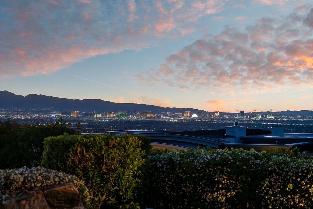 This elevated exterior view captures a breathtaking panoramic vista of the Las Vegas city skyline at dusk, framed by lush foreground landscaping. The scene features a modern roofline in the immediate foreground, leading the eye toward the sprawling urban lights and distant mountain silhouettes under a vibrant, cloud-streaked sunset sky. The perspective emphasizes the property's prime location and dramatic, high-altitude vantage point.