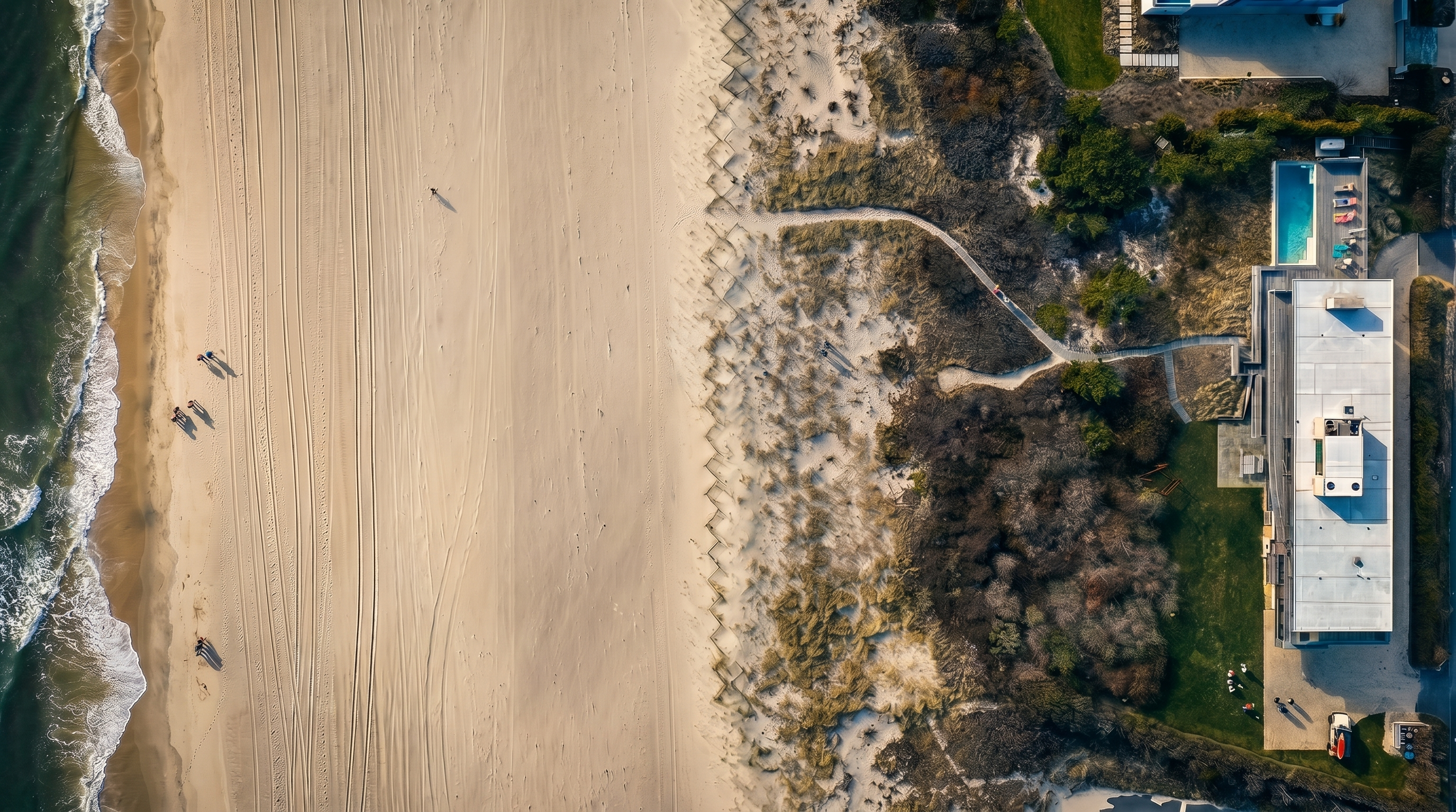 This high-altitude aerial perspective captures a modern, linear residence situated directly along a pristine sandy coastline. The property features a rectangular swimming pool, a manicured lawn, and a private path winding through natural dune vegetation toward the beach. The clean, minimalist architecture of the home contrasts with the organic textures of the surrounding landscape, emphasizing its secluded and exclusive beachfront location.