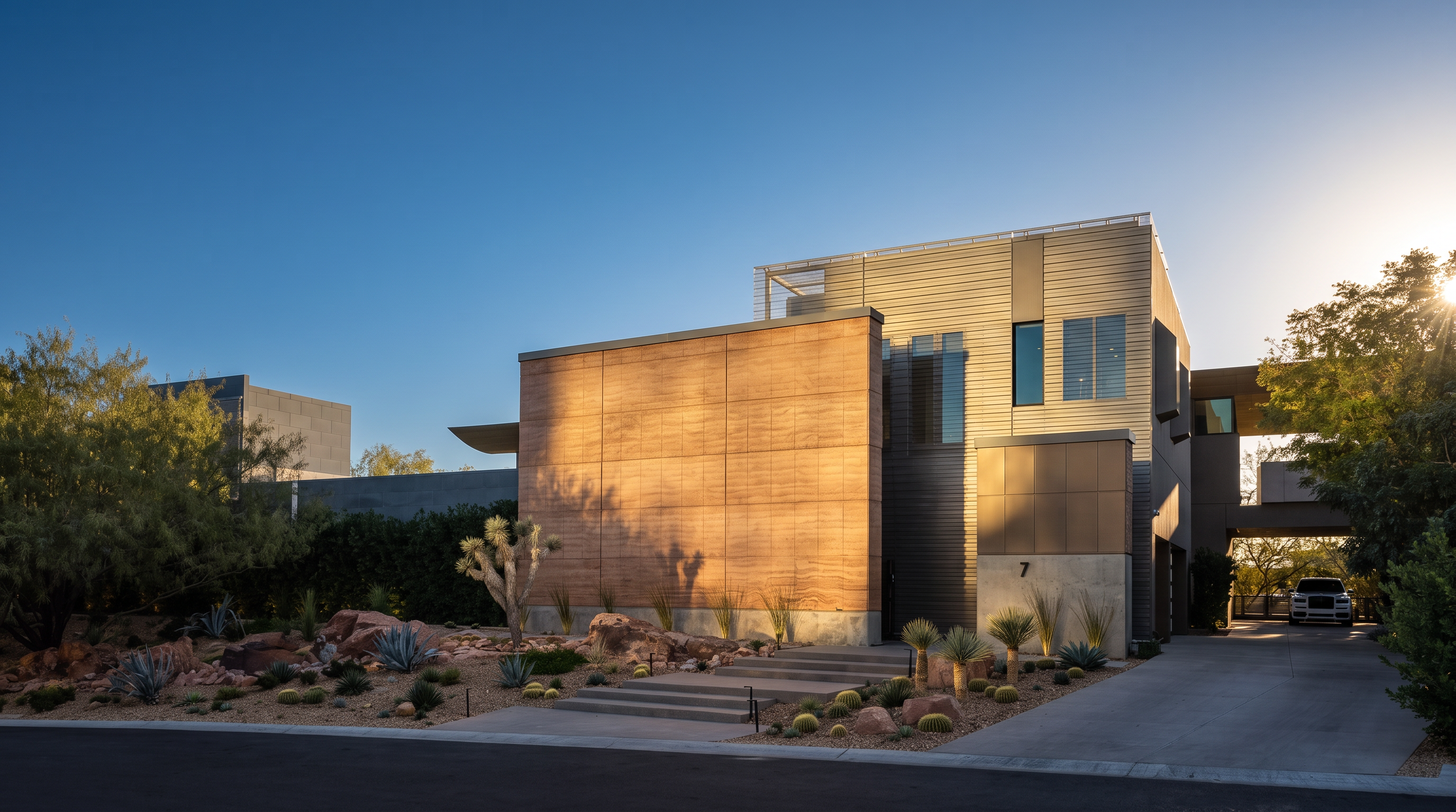 This striking modern residence features a bold architectural design with a prominent, textured stone-clad facade and clean, geometric lines. The front entryway is accessed by a set of wide concrete steps, flanked by desert-inspired landscaping with cacti and boulders illuminated by warm uplighting. A paved driveway leads to a side-entry garage, while the dusk lighting creates an inviting and sophisticated atmosphere.