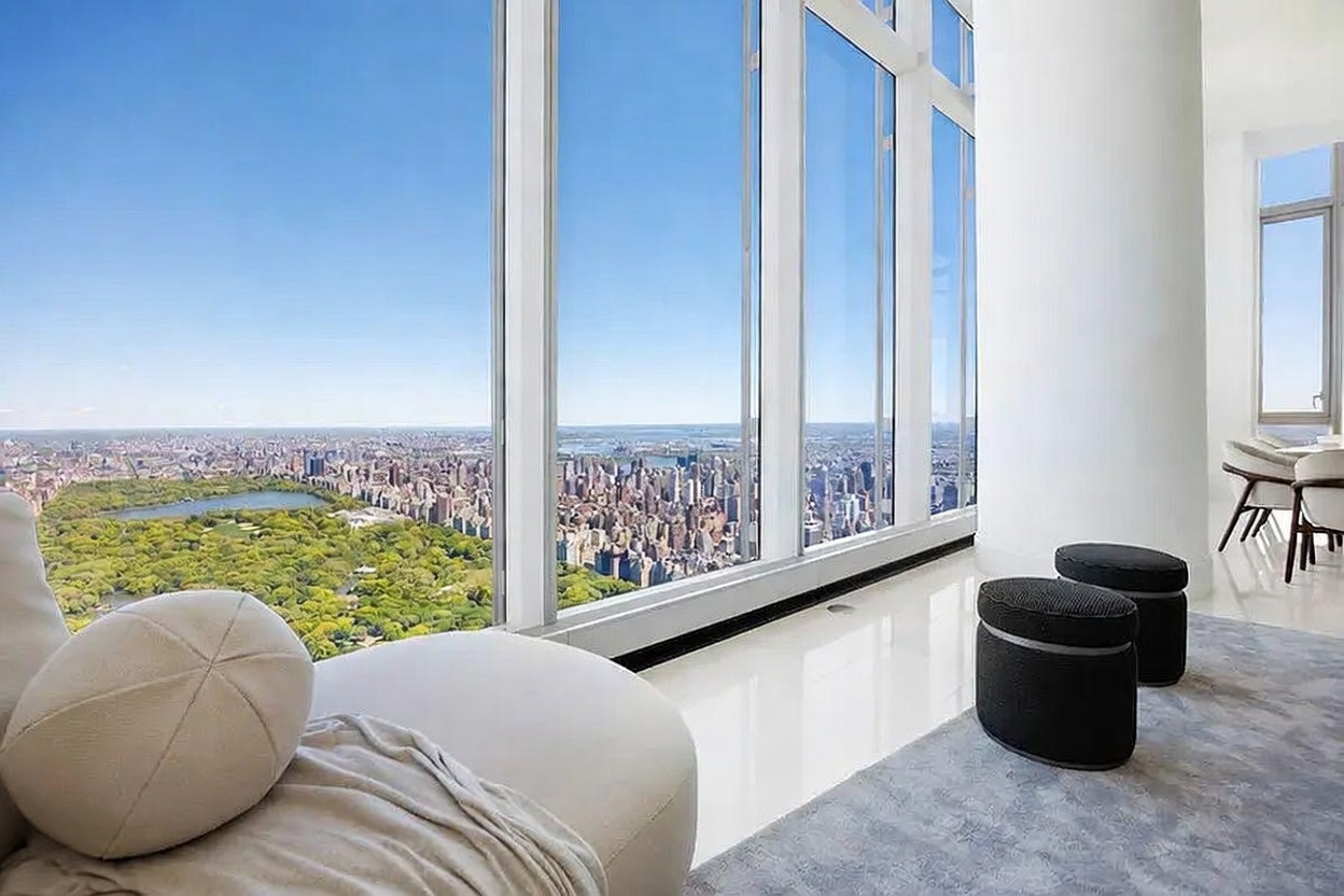 This high-rise living space features expansive floor-to-ceiling windows that offer a panoramic view of a city park and skyline. The room is furnished with a plush, light-colored lounge chair in the foreground and two dark, cylindrical ottomans resting on a grey area rug. The minimalist interior design emphasizes the dramatic urban vista, creating a bright and airy atmosphere.