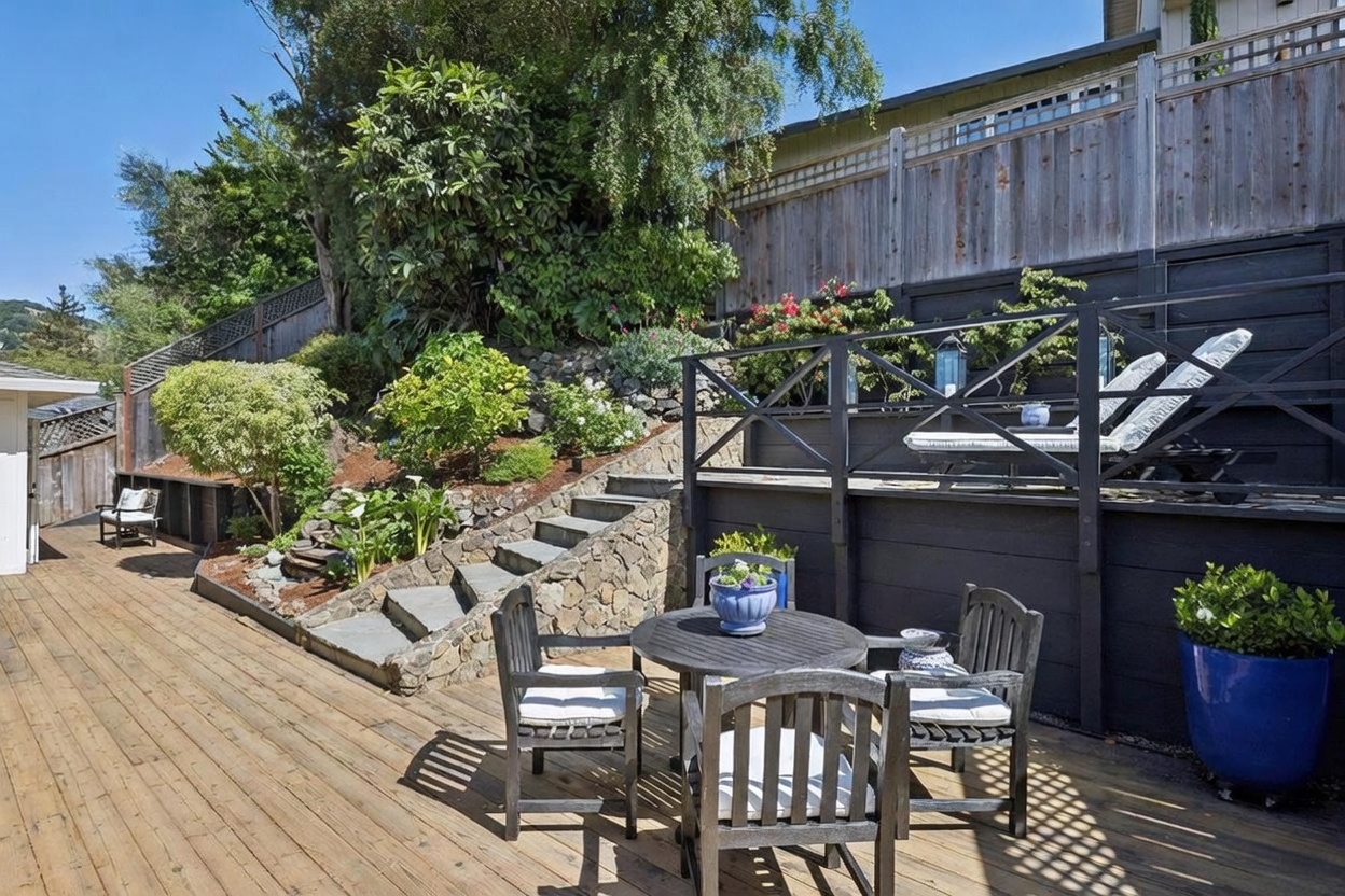 This inviting outdoor living space features a multi-level wooden deck with a dining set and a raised lounge area equipped with sun loungers. A stone-stepped staircase leads up to a lush, terraced garden area, framed by a dark-painted wooden fence and mature trees. The perspective captures a serene, private backyard retreat perfect for relaxation and entertaining.