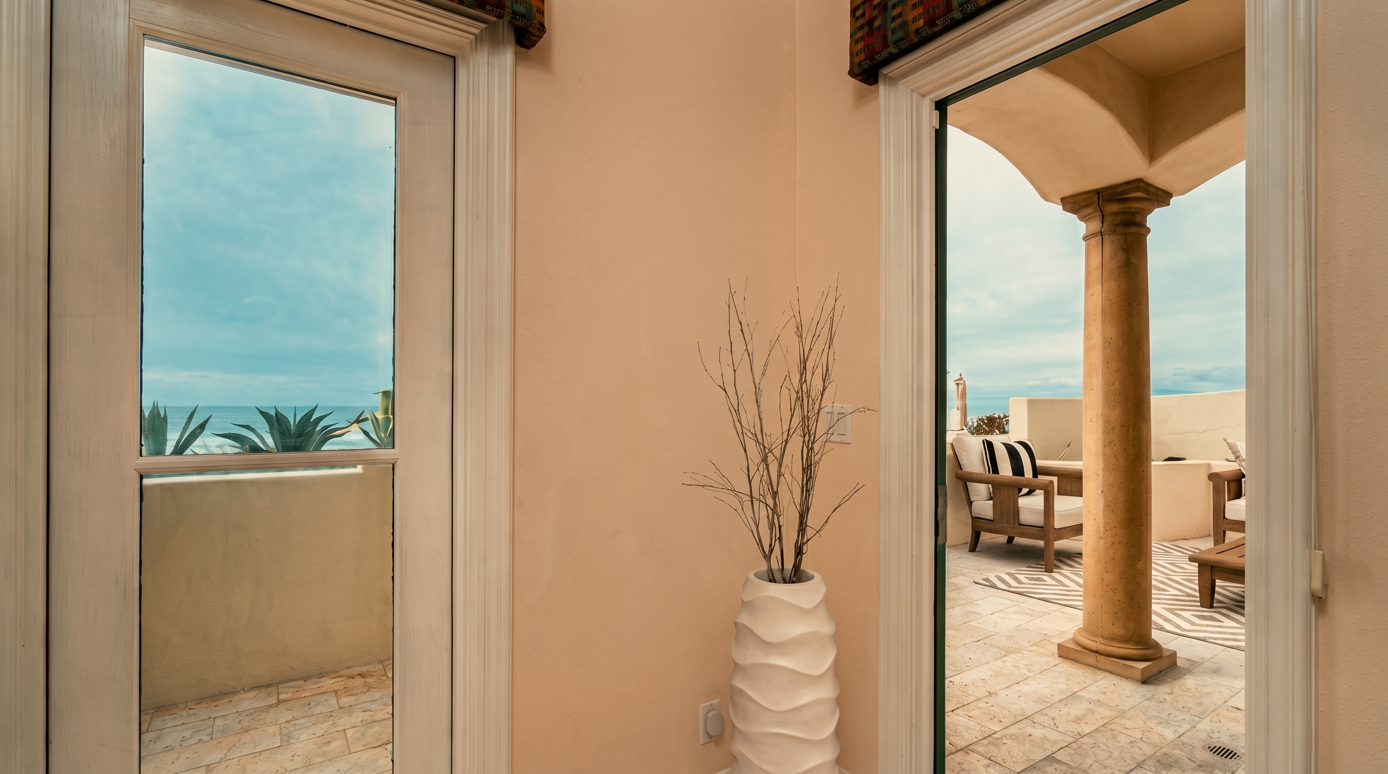 This interior space serves as a transition area, featuring a tall white decorative vase with dried branches positioned between two glass doors. The doors open onto a tiled balcony area that offers an expansive view of the ocean and a covered patio space with outdoor furniture. The neutral wall color and clean lines create a bright, airy atmosphere that emphasizes the connection between the indoor and outdoor living spaces.
