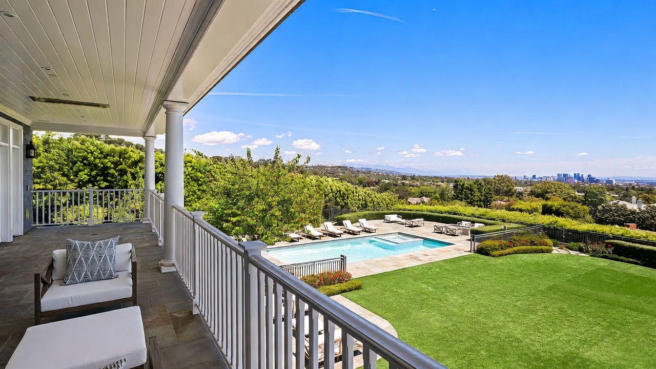 This elevated balcony offers a sweeping, panoramic view of a lush backyard, a sparkling swimming pool, and a distant cityscape under a clear blue sky. The covered deck features stone flooring and comfortable outdoor seating, providing a seamless transition to the expansive lawn and pool area below. The perspective is captured from a high vantage point, emphasizing the property's prime location and serene outdoor living space.