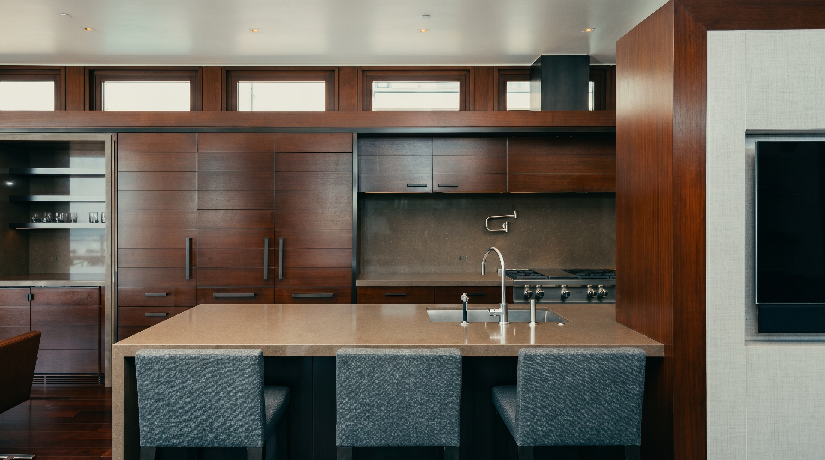 This modern kitchen features dark wood cabinetry and a large central island with a stone countertop and three gray upholstered bar stools. The space is equipped with a professional-grade range and a sleek faucet, set against a backdrop of horizontal windows that allow natural light to filter in. The overall aesthetic is sophisticated and minimalist, emphasizing clean lines and a rich, warm color palette.