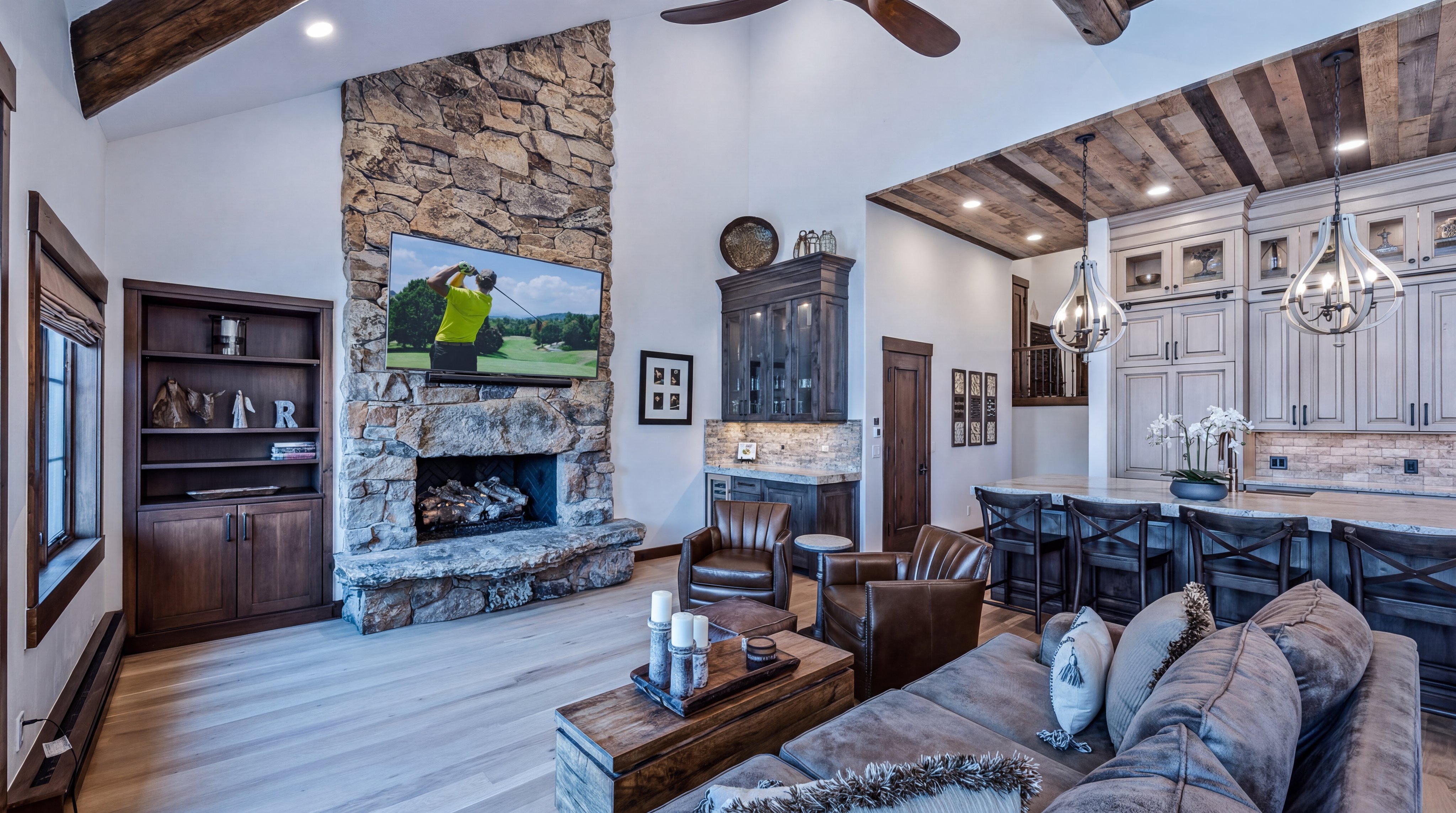 This grand living room features a striking floor-to-ceiling stone fireplace with an integrated television that serves as the room's focal point. The open-concept design flows seamlessly into an adjacent kitchen area, showcasing rustic elements like reclaimed wood ceilings and leather furniture. High vaulted ceilings and rich wood cabinetry create a warm, inviting, and sophisticated atmosphere.