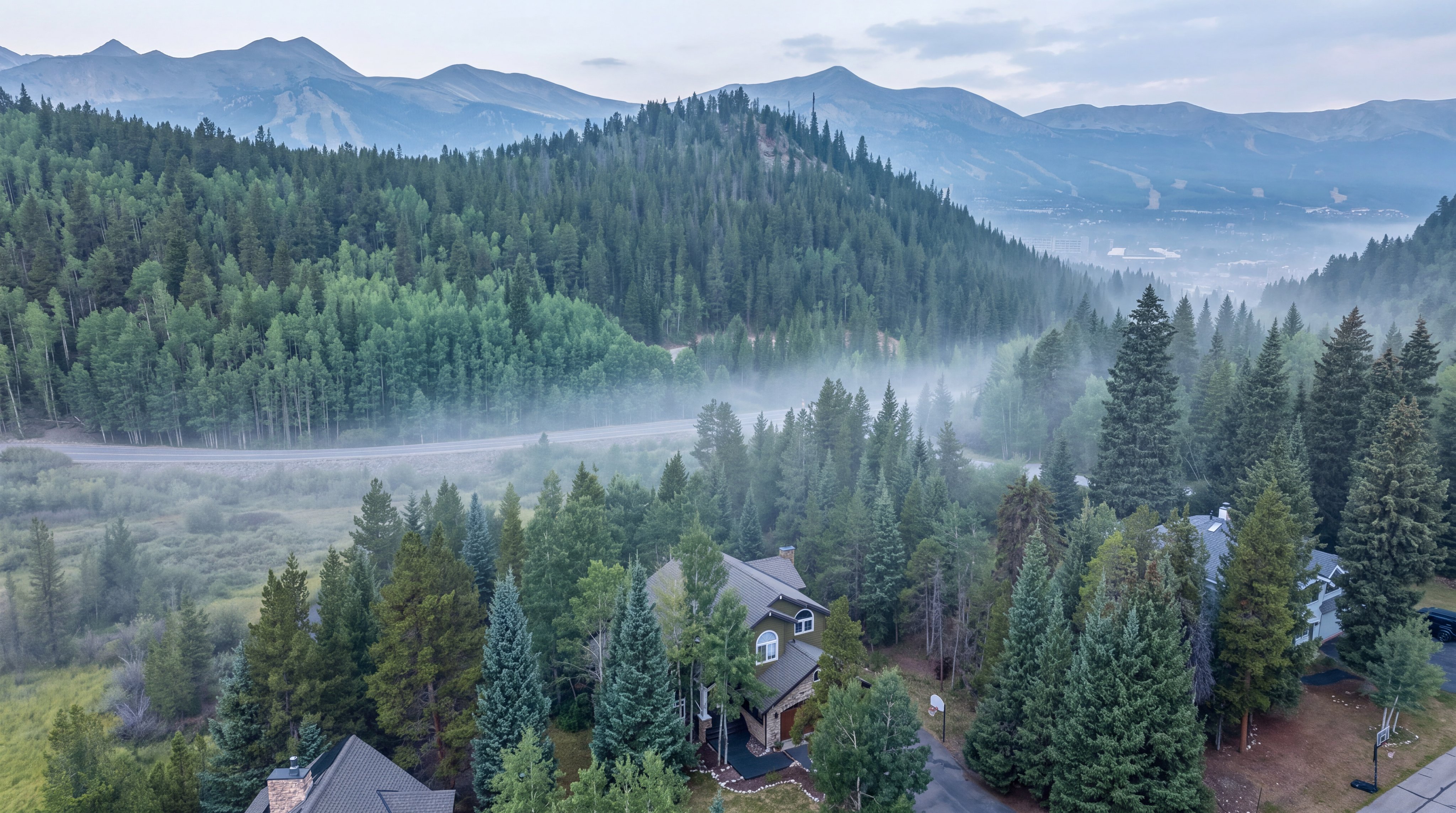This aerial view captures a scenic, multi-level mountain home nestled within a dense evergreen forest. The property features a classic shingled roof and a secluded driveway, set against a breathtaking backdrop of misty peaks and mountain vistas. The atmosphere is serene and private, offering a quintessential high-altitude retreat.