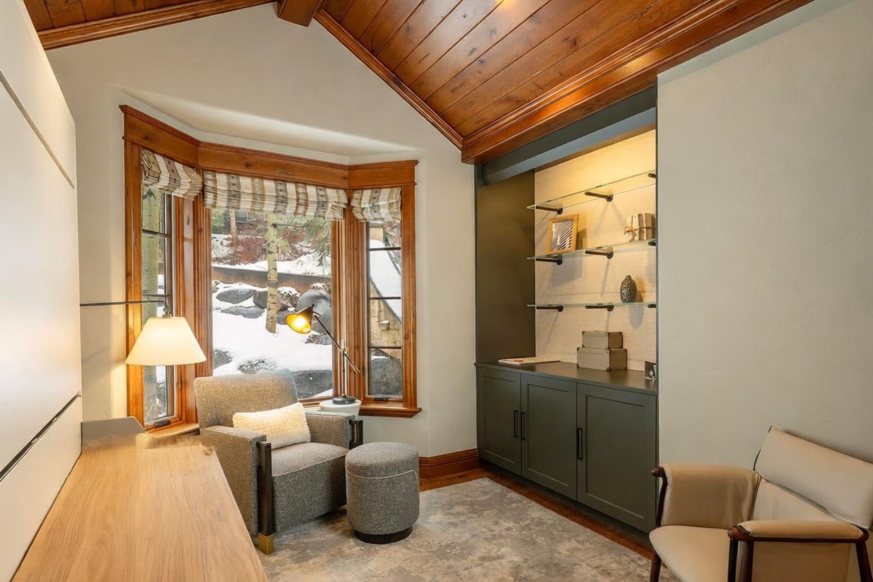 This cozy home office features a vaulted wood-paneled ceiling and a large bay window that provides a view of a snowy outdoor setting. The room is furnished with a comfortable armchair and ottoman, a desk, and a built-in shelving unit with glass display shelves and dark cabinetry. The space is illuminated by a desk lamp and natural light, creating a warm and inviting atmosphere for work or relaxation.
