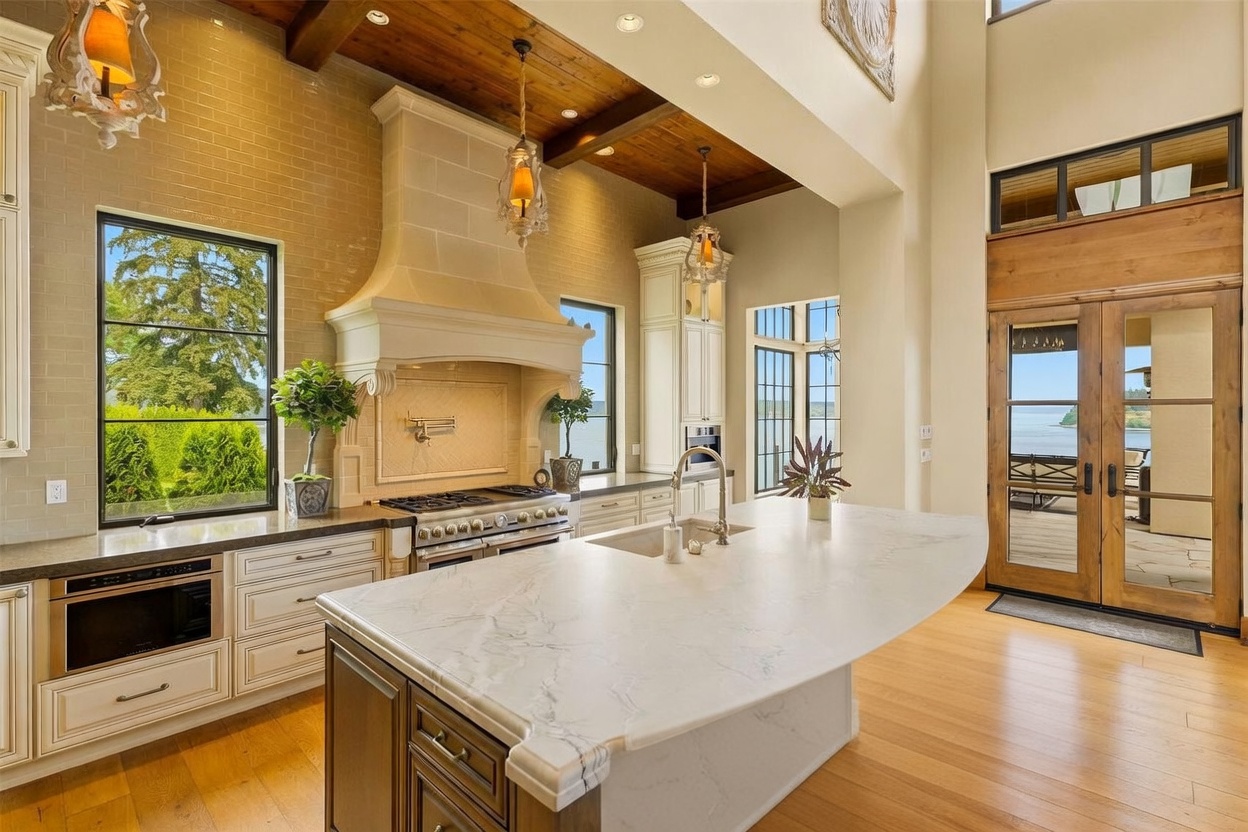 This elegant kitchen features a large central island with a stone countertop and seating, complemented by custom cabinetry and a prominent range hood. The space is defined by a high, wood-beamed ceiling and large windows that provide expansive views of the water outside. The combination of warm wood tones, neutral cabinetry, and sophisticated lighting creates a luxurious and inviting atmosphere.