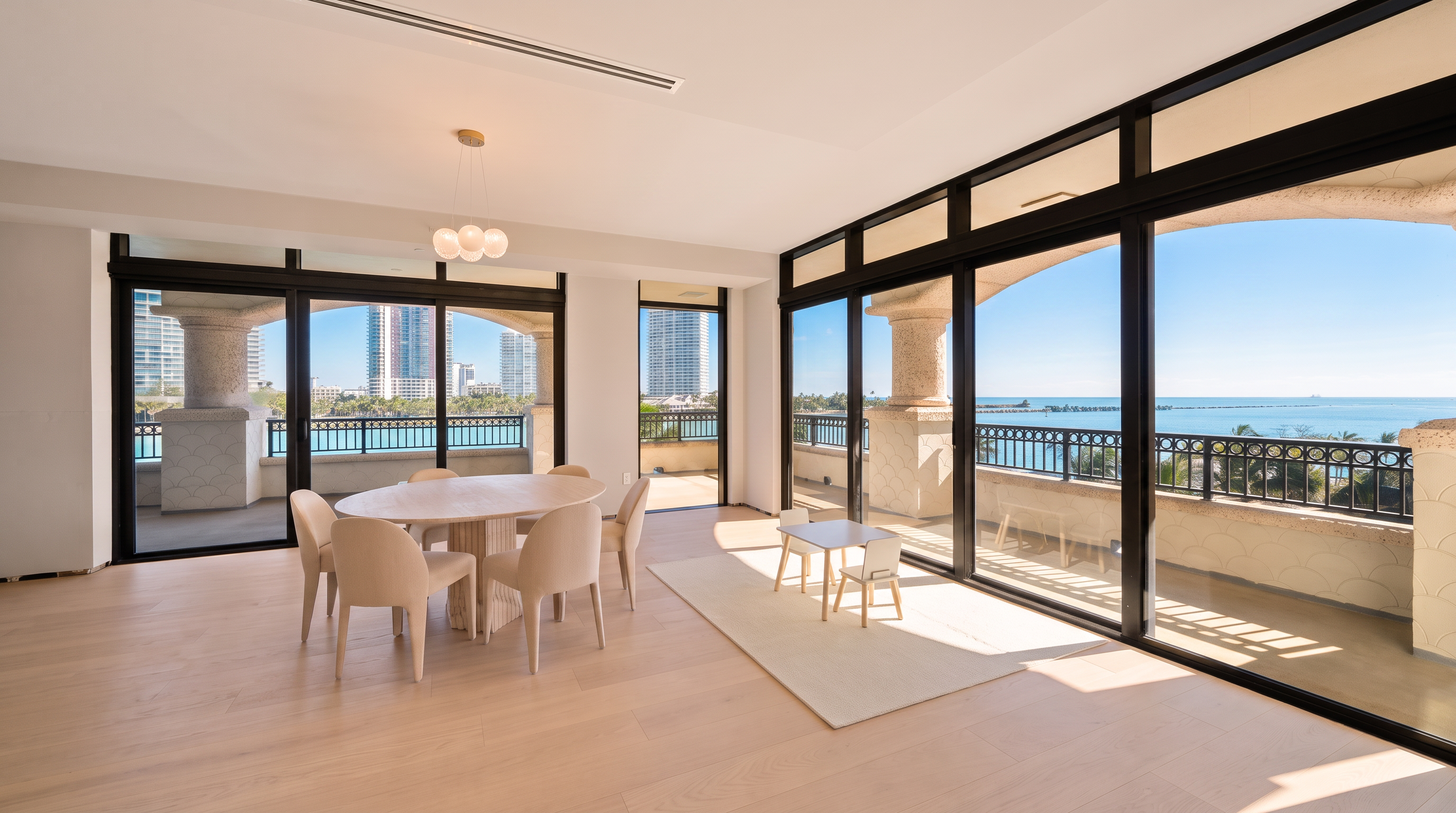 This bright and airy dining area features light wood flooring and a neutral color palette, centered around a round dining table with matching chairs. Large glass sliding doors provide expansive views of the ocean and city skyline, flooding the space with natural light. The room is minimalist in design, emphasizing the seamless transition between the indoor living space and the outdoor balcony.