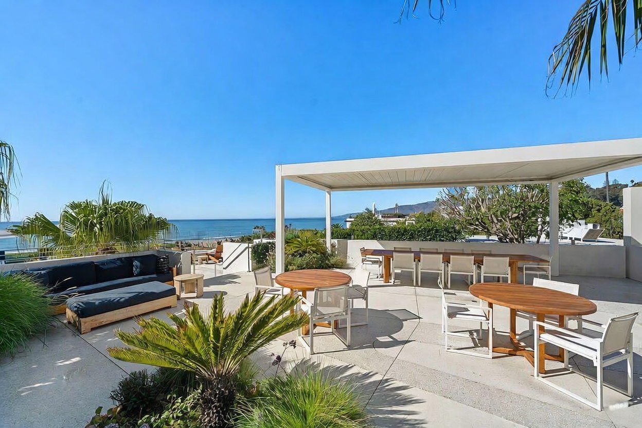 This expansive, sun-drenched outdoor terrace offers a sophisticated coastal living experience with panoramic ocean views. The space features a covered dining area with a long wooden table, a separate circular dining set, and a comfortable lounge area with dark-cushioned seating. Lush landscaping, including palm-like plants, adds a touch of greenery to the concrete patio, creating a serene and luxurious atmosphere perfect for entertaining.