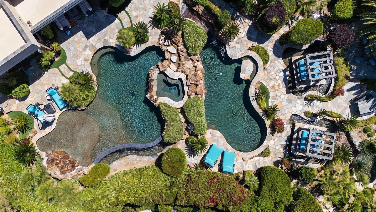 This high-angle aerial view captures a luxurious, resort-style backyard featuring a large, free-form swimming pool with a central rock-encased spa. The pool area is surrounded by natural stone decking, lush landscaping, and two shaded cabanas with blue lounge seating. The scene conveys a serene, private oasis perfect for outdoor entertaining and relaxation.
