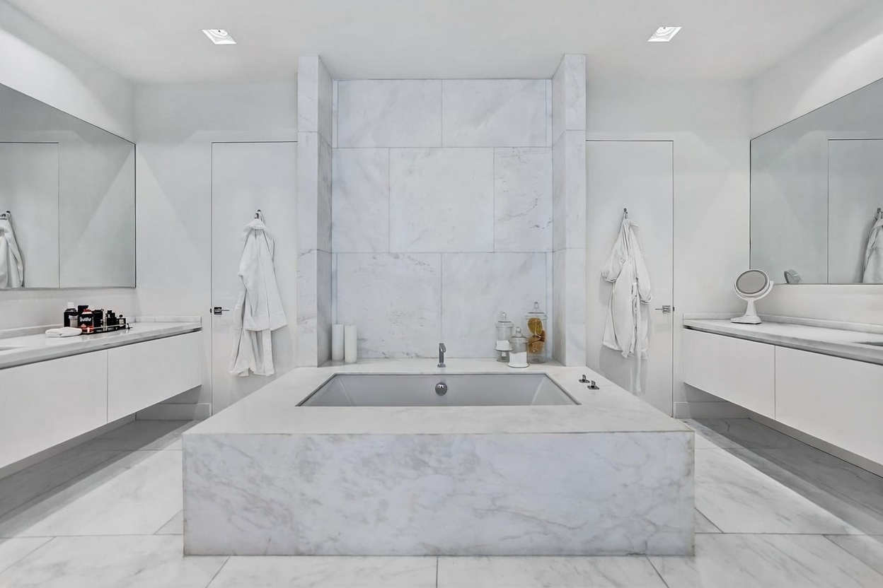 This luxurious primary bathroom features a central, stone-clad soaking tub as the focal point, set against a wall of large-format stone tiles. The symmetrical layout includes dual vanities with expansive mirrors on either side, flanked by white doors and hanging robes. The space is finished in a bright, monochromatic palette that creates a clean, spa-like atmosphere.