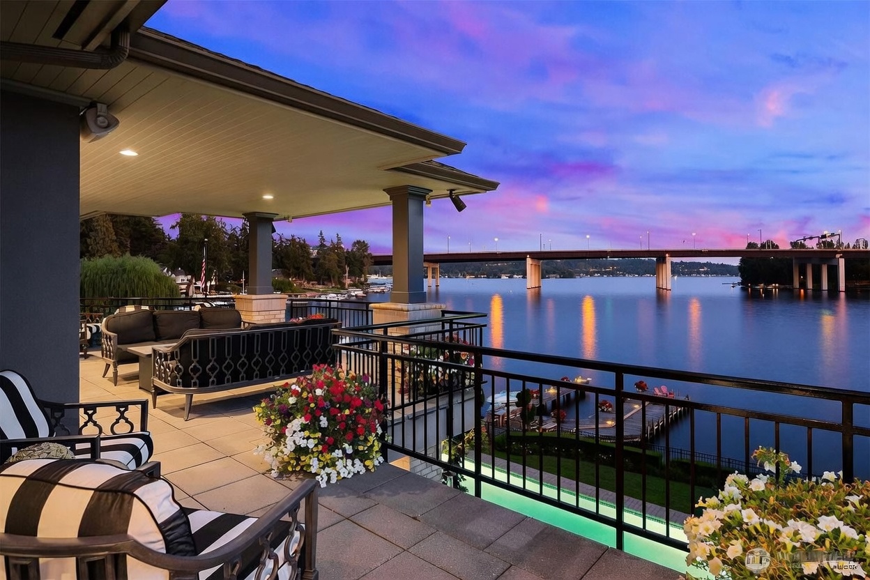 This expansive covered balcony offers a luxurious outdoor living space with comfortable lounge seating and vibrant floral arrangements. The space overlooks a serene lake at twilight, featuring a prominent bridge in the distance and a glowing pool visible below the railing. The scene captures a sophisticated, tranquil atmosphere perfect for evening relaxation.