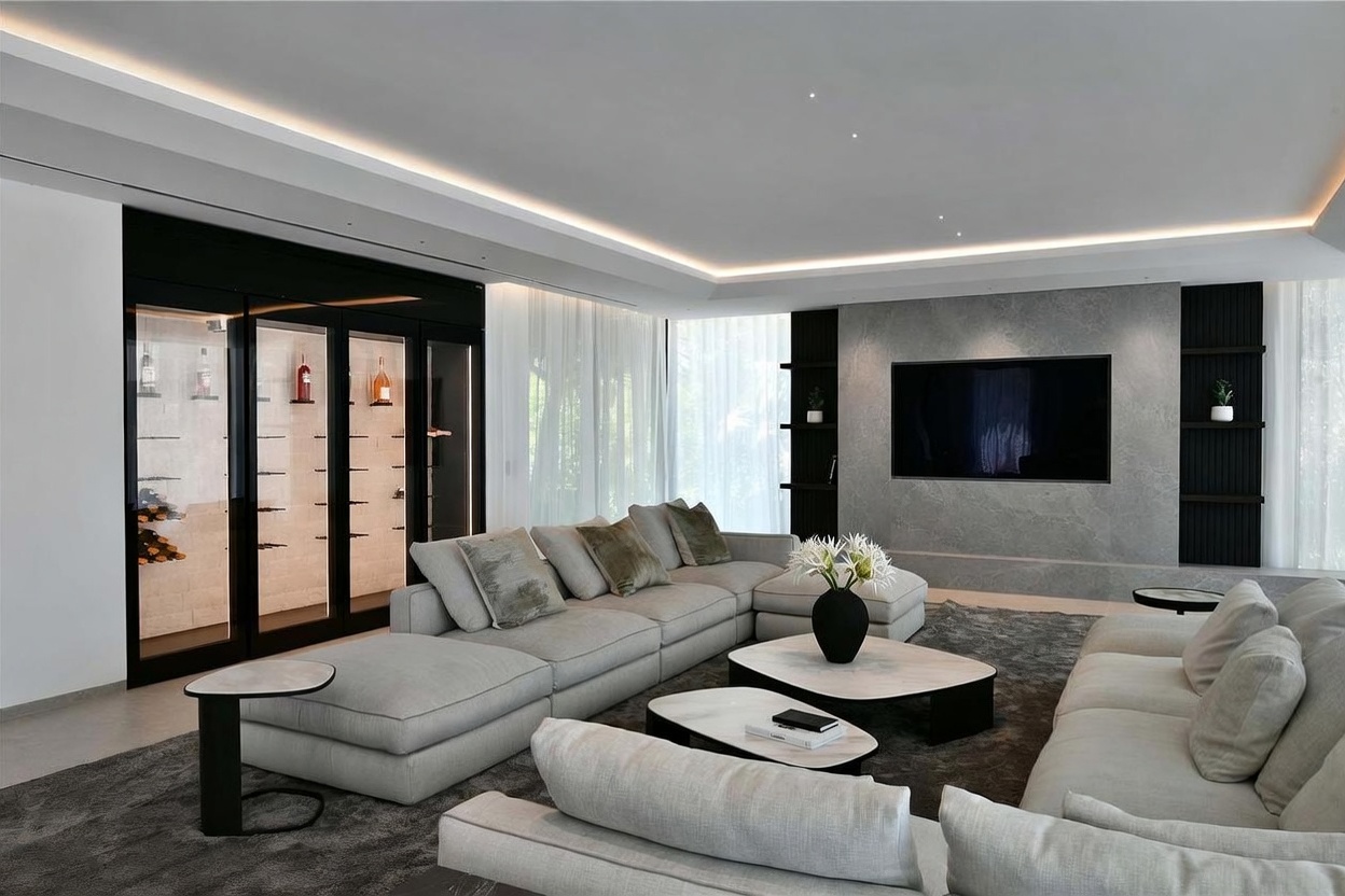 This sophisticated living room features a large, light-toned sectional sofa arranged around two nested coffee tables on a plush, dark area rug. A prominent stone-clad wall serves as the focal point, housing a wall-mounted television and integrated shelving, while a sleek, glass-enclosed wine display adds a touch of luxury to the side. The space is illuminated by recessed lighting and soft, ambient cove lighting, creating an inviting and modern atmosphere.