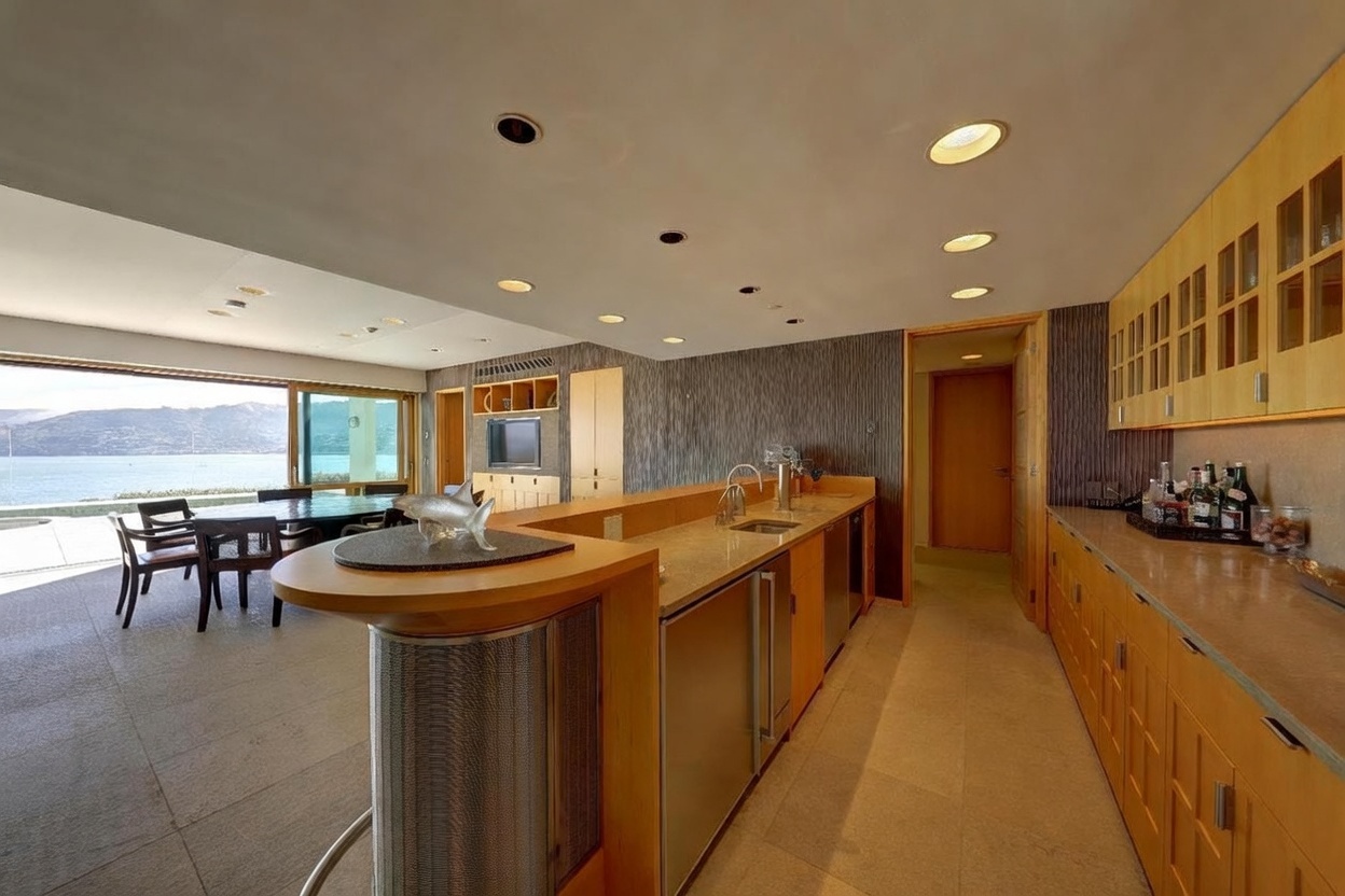 This modern kitchen features a long island with a curved breakfast bar and a built-in sink, complemented by light wood cabinetry and stone countertops. The space flows seamlessly into an adjacent dining area that opens to a scenic waterfront view through large glass sliding doors. The room is illuminated by numerous recessed lights, creating a bright and airy atmosphere with a contemporary, open-concept design.