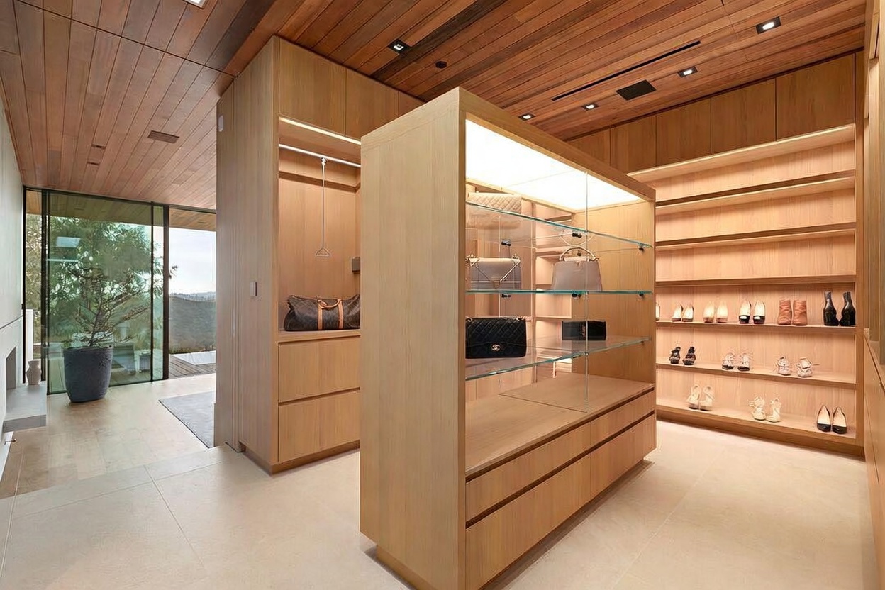 This luxurious, modern walk-in closet features warm wood paneling on the walls and ceiling, creating a sophisticated and cohesive aesthetic. The space is anchored by a central illuminated display island with glass shelving for handbags, complemented by extensive open shelving for footwear and hanging storage. A large floor-to-ceiling glass wall provides a seamless transition to an outdoor area, bathing the room in natural light and enhancing the open, airy atmosphere.