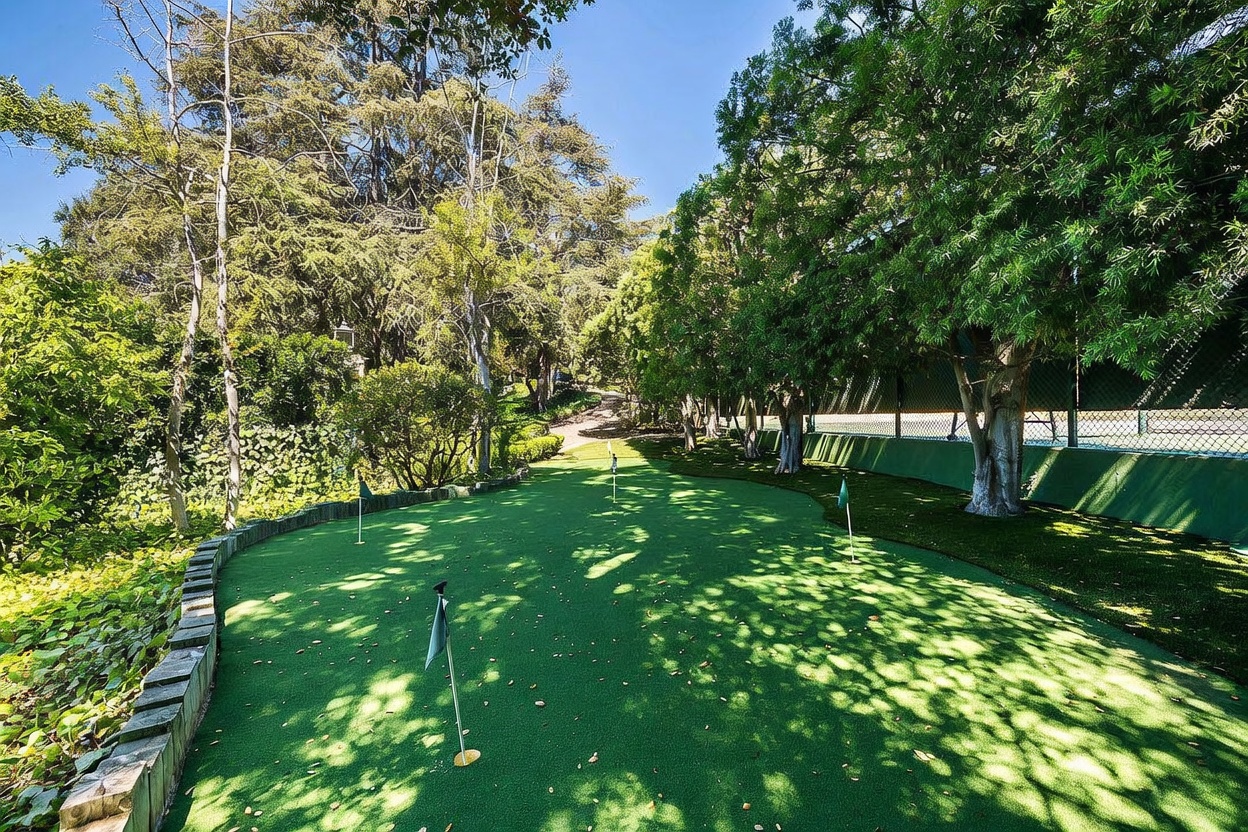 This backyard features a private putting green with multiple holes, set against a backdrop of mature trees and lush greenery. A stone retaining wall borders the putting area, while a green fence separates the space from an adjacent tennis court. The scene is captured from a low, eye-level perspective, emphasizing the well-manicured lawn and the tranquil, shaded atmosphere of the outdoor space.