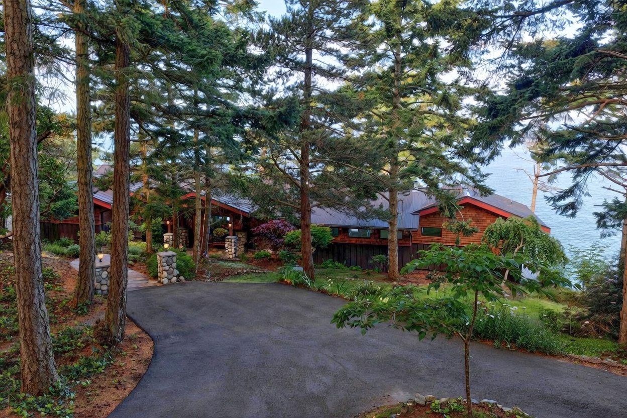 This elevated perspective captures a secluded, wood-shingled home nestled within a dense forest of tall evergreen trees. The house features a multi-gabled roofline, stone-accented pillars at the entryway, and a winding paved driveway leading to the front entrance. The setting offers a serene, natural atmosphere with a glimpse of the nearby water visible through the trees.