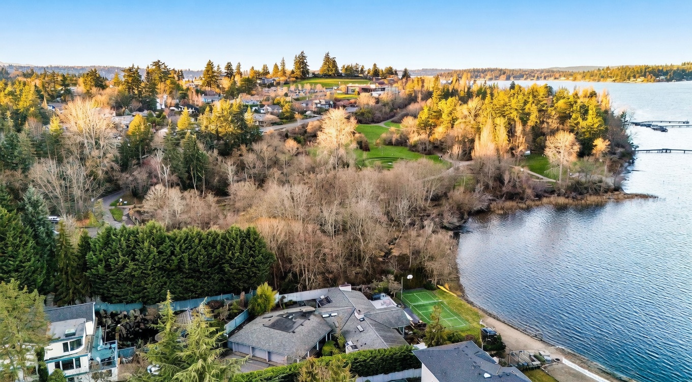 This high-angle aerial view captures a waterfront property nestled among lush, mature trees and greenery. The home features a multi-pitched roofline, a private tennis court, and a long wooden dock extending into the calm lake. The surrounding landscape includes a sprawling park-like area, highlighting the serene and secluded nature of this lakeside estate.
