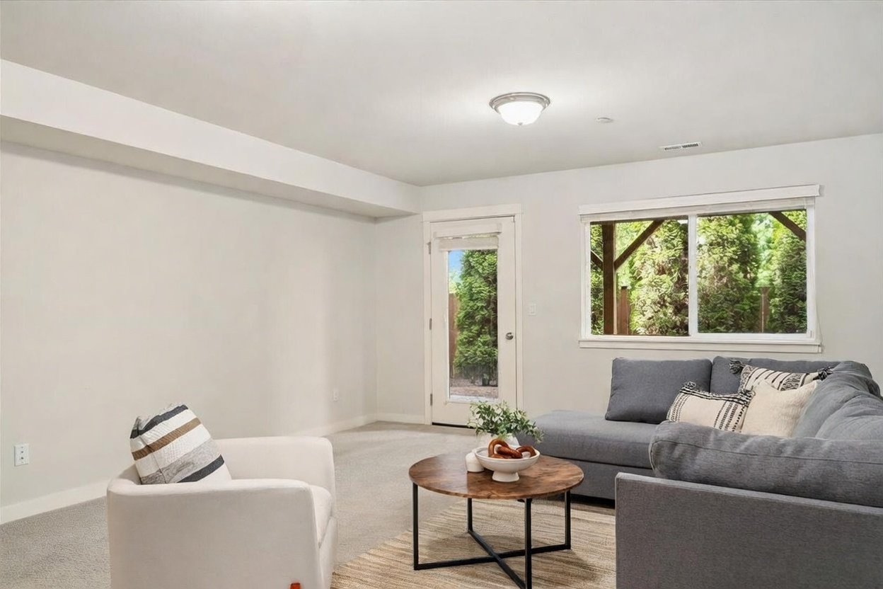 This bright and inviting living room features neutral-toned walls and plush carpeting, creating a comfortable atmosphere. The space is furnished with a large grey sectional sofa and a modern white armchair, centered around a round wooden coffee table. A glass-paned door and a wide window provide natural light and a view of the lush greenery outside.