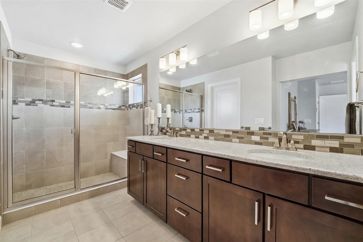 This primary bathroom features a double vanity with dark wood cabinetry, a light-colored stone countertop, and a large wall-to-wall mirror. To the left, a glass-enclosed walk-in shower with neutral-toned wall tiles and a built-in bench provides a clean, modern aesthetic. The space is well-lit by overhead vanity lighting and recessed ceiling lights, creating a bright and functional atmosphere.