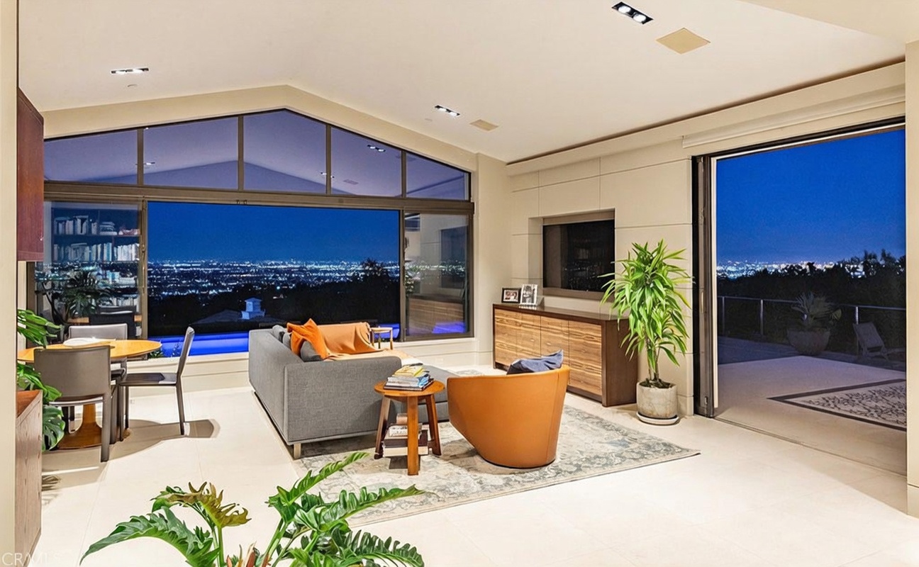 This modern, open-concept living area features a neutral color palette and expansive glass walls that frame a stunning panoramic view of a city at night. The space is furnished with a comfortable gray sofa, a warm-toned leather armchair, and a wood-finished media console, all arranged on a patterned area rug. A dining table is positioned to the left, while large sliding glass doors open to an outdoor patio, creating a seamless transition between indoor and outdoor living.