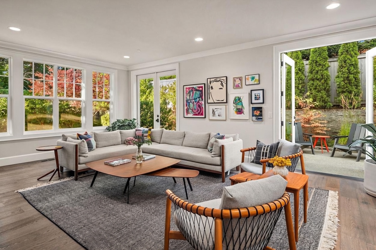 This bright and airy living room features a large, light-colored sectional sofa arranged around a tiered wooden coffee table on a textured area rug. The space is accented by two mid-century modern style chairs, a gallery wall of colorful artwork, and expansive windows that provide ample natural light. Large glass doors open directly onto a private patio, creating a seamless transition between the indoor living area and the outdoor garden space.
