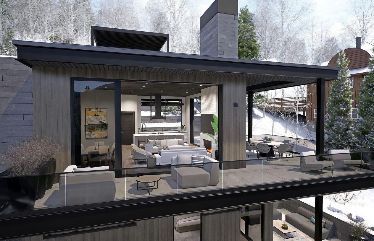 This modern mountain residence features a seamless transition between indoor and outdoor living spaces, highlighted by a spacious balcony with glass railings. The open-concept interior showcases a contemporary living area and kitchen, while the covered deck provides an ideal setting for al fresco dining amidst a snowy, wooded landscape. The architectural design emphasizes clean lines, dark wood-toned siding, and a prominent stone chimney breast that anchors the structure.