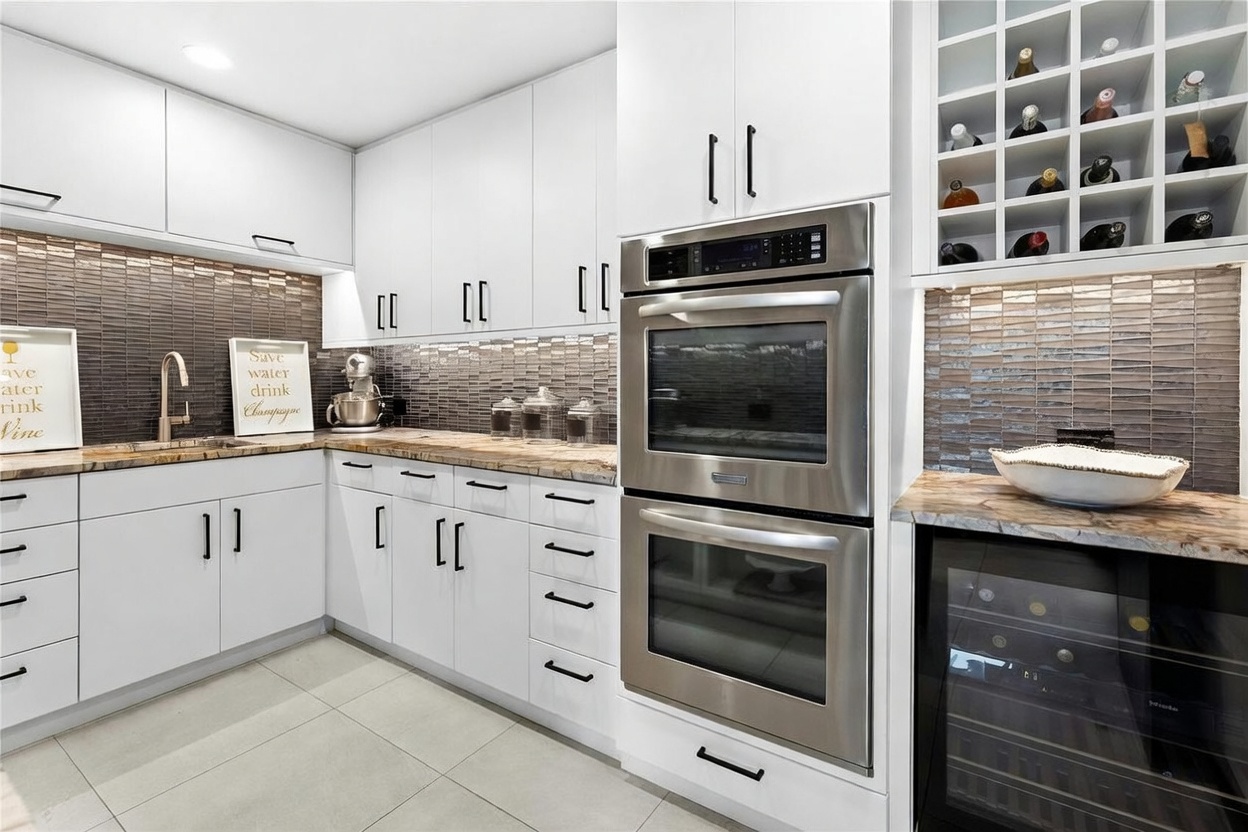 This modern, high-end kitchen features sleek white cabinetry, stainless steel double wall ovens, and a built-in wine refrigerator. The space is accented by a textured dark tile backsplash, marble-patterned countertops, and a dedicated wine rack, creating a sophisticated and functional culinary environment. The perspective is a wide-angle shot capturing the efficient layout and premium finishes of this luxury home space.
